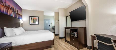 A hotel room with a bed, a desk, a chair, a TV, and a bathroom in the background.