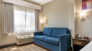 A hotel room with a blue sofa, a bedside table with a lamp, a heater, and a painting on the wall.