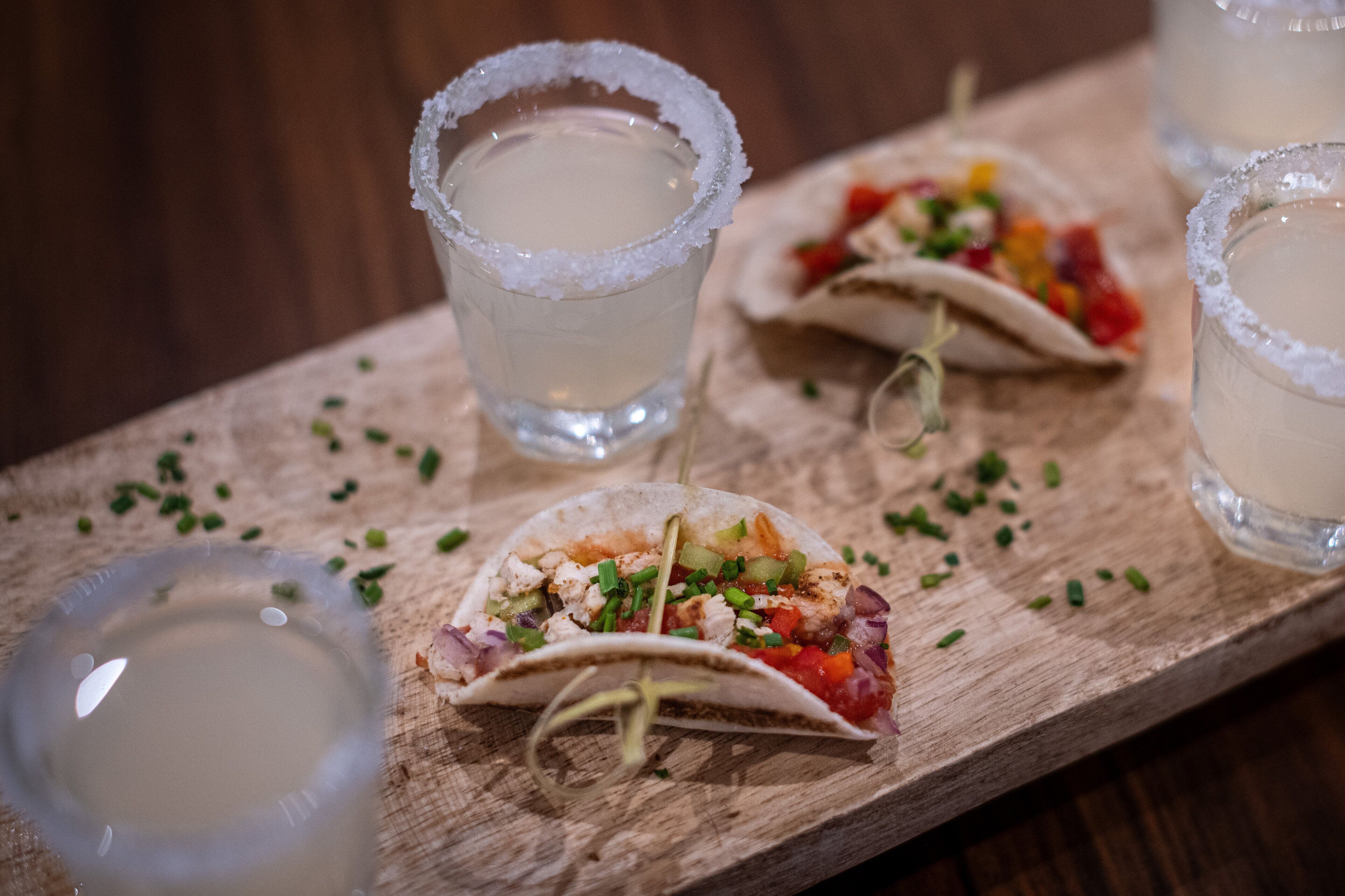 A wooden serving board with three tacos and two glasses of a clear beverage, garnished with a rim of salt.