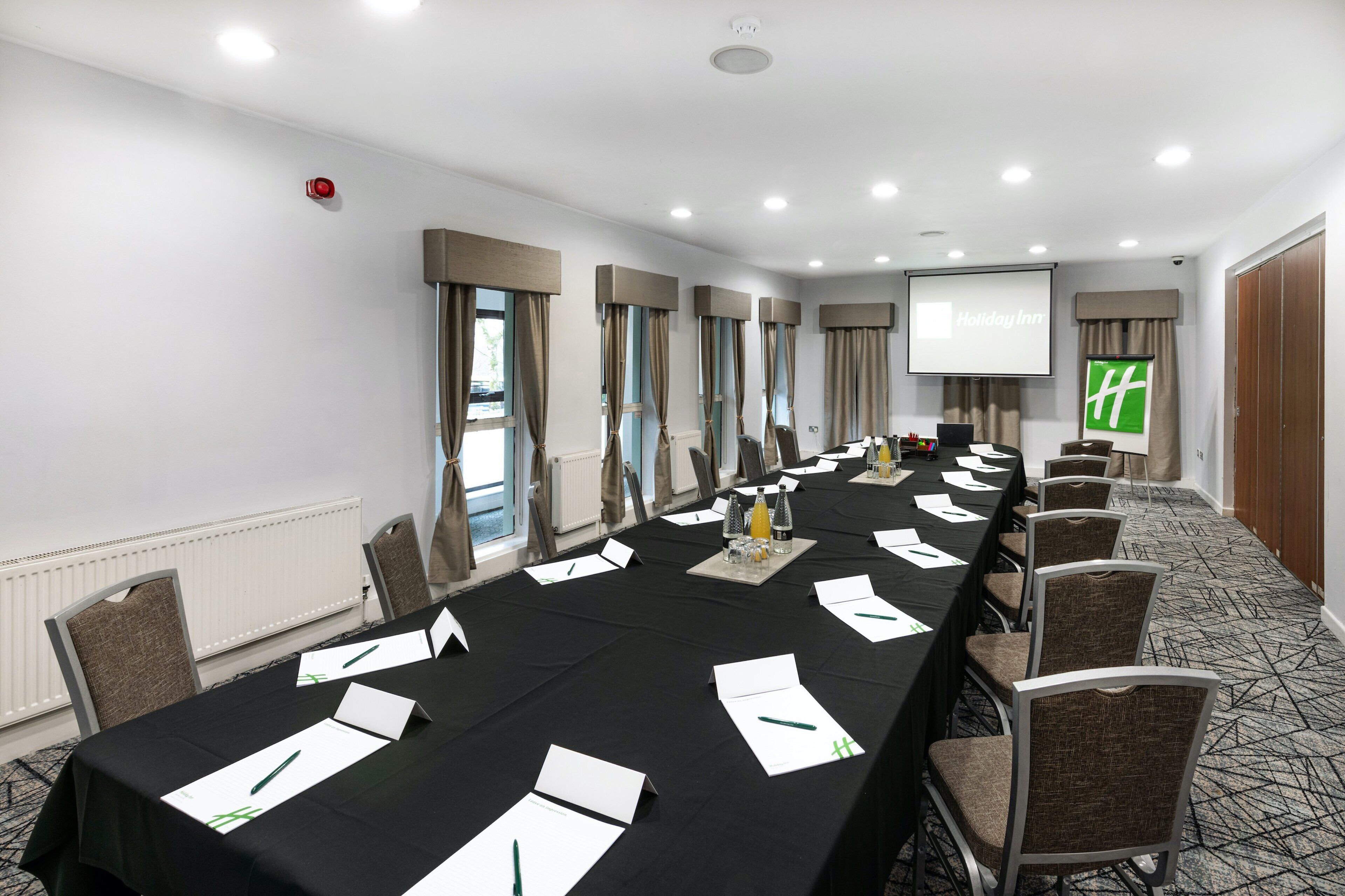 A conference room with a long table, chairs, a projector screen, and a screen displaying 'Holiday Inn'.