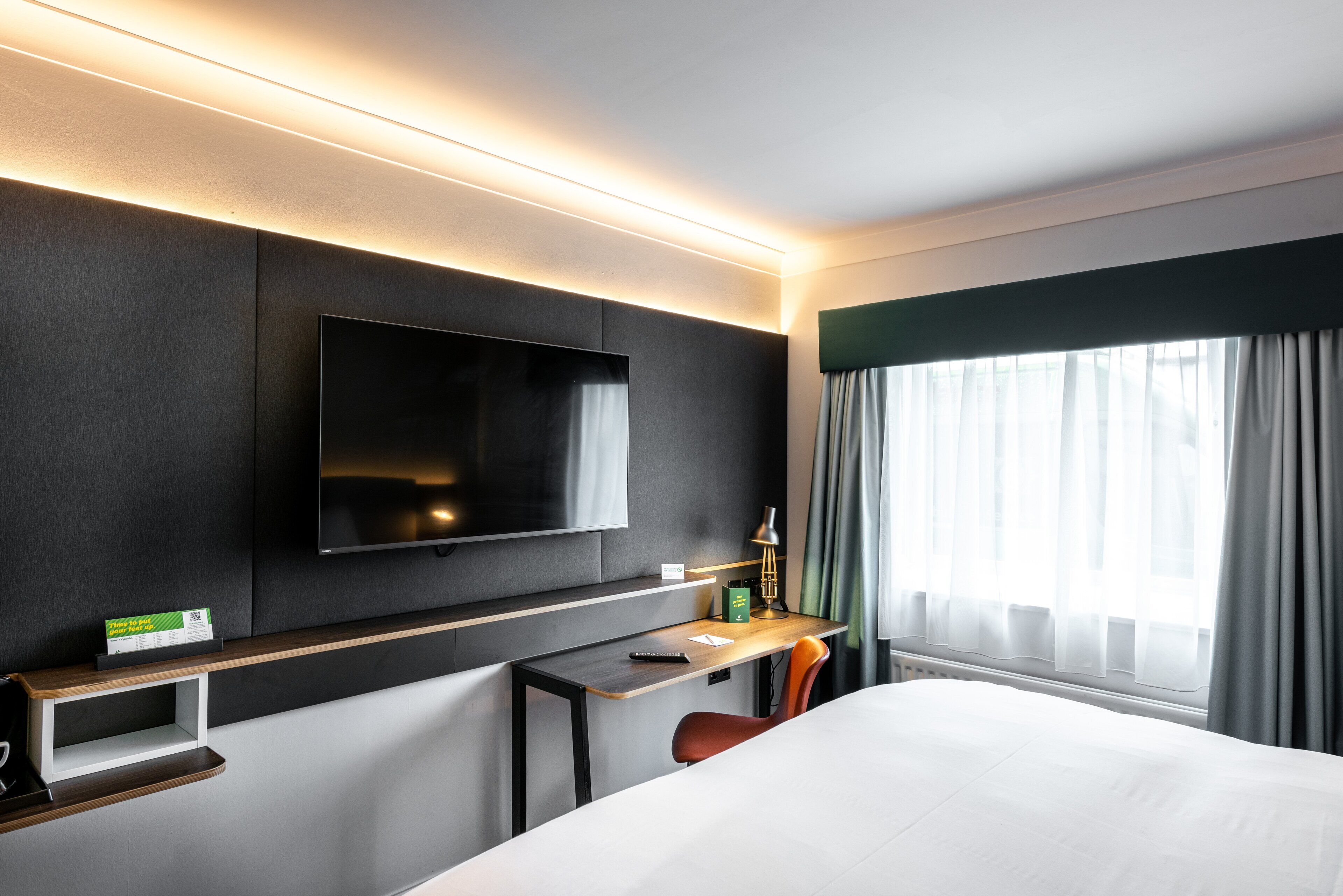 A modern hotel room with a large bed, a flat-screen TV mounted on the wall, a desk with a chair, and a window with curtains.