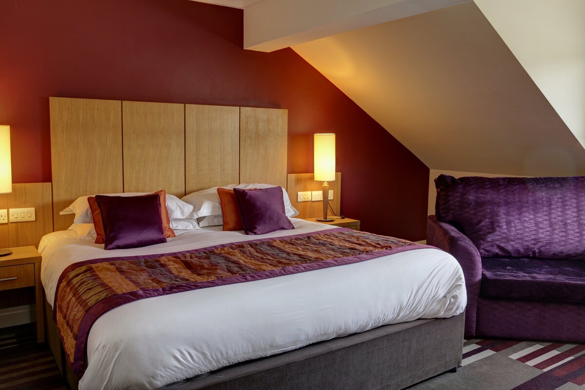 Una habitación de hotel con una cama grande, dos lámparas de noche, un sillón morado y un cabecero de madera.
