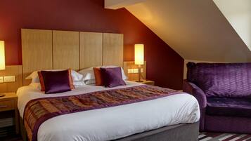 Habitación de hotel con una cama grande, dos lámparas de noche, un cabecero de madera, un sillón púrpura y una manta estampada.