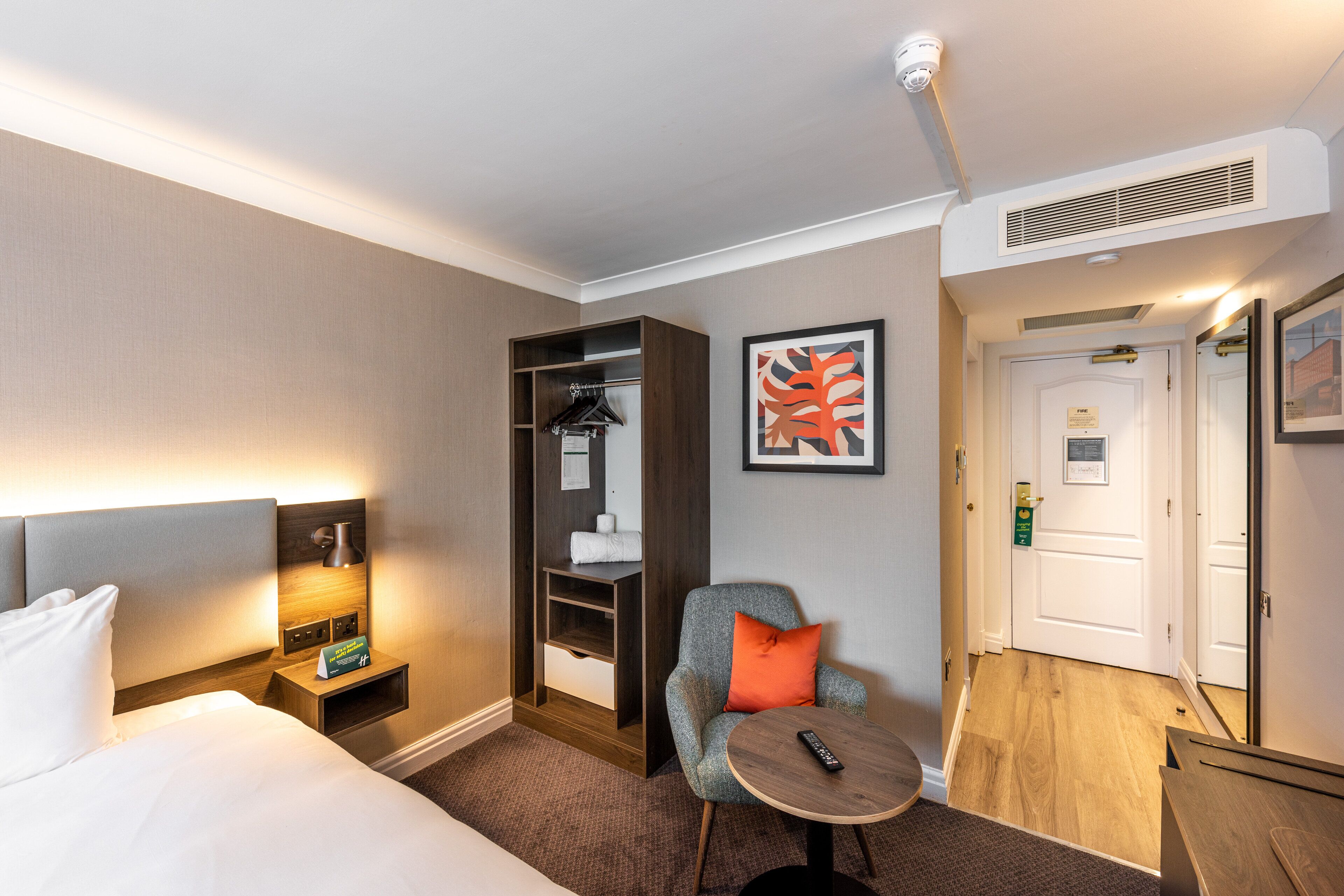 Ein Hotelzimmer mit einem Bett, einem Nachttisch mit Lampe, einem Sessel mit Kissen, einem kleinen Tisch mit Fernbedienung, einem Kleiderschrank und einer Tür.