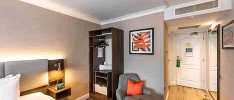Una habitación de hotel con una cama, una mesita de noche con una lámpara, una silla con un cojín, una pequeña mesa con un control remoto, un armario y una puerta.