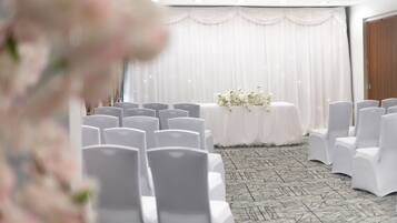 A conference room with rows of white chairs, a table with white flowers, and a white curtain backdrop.