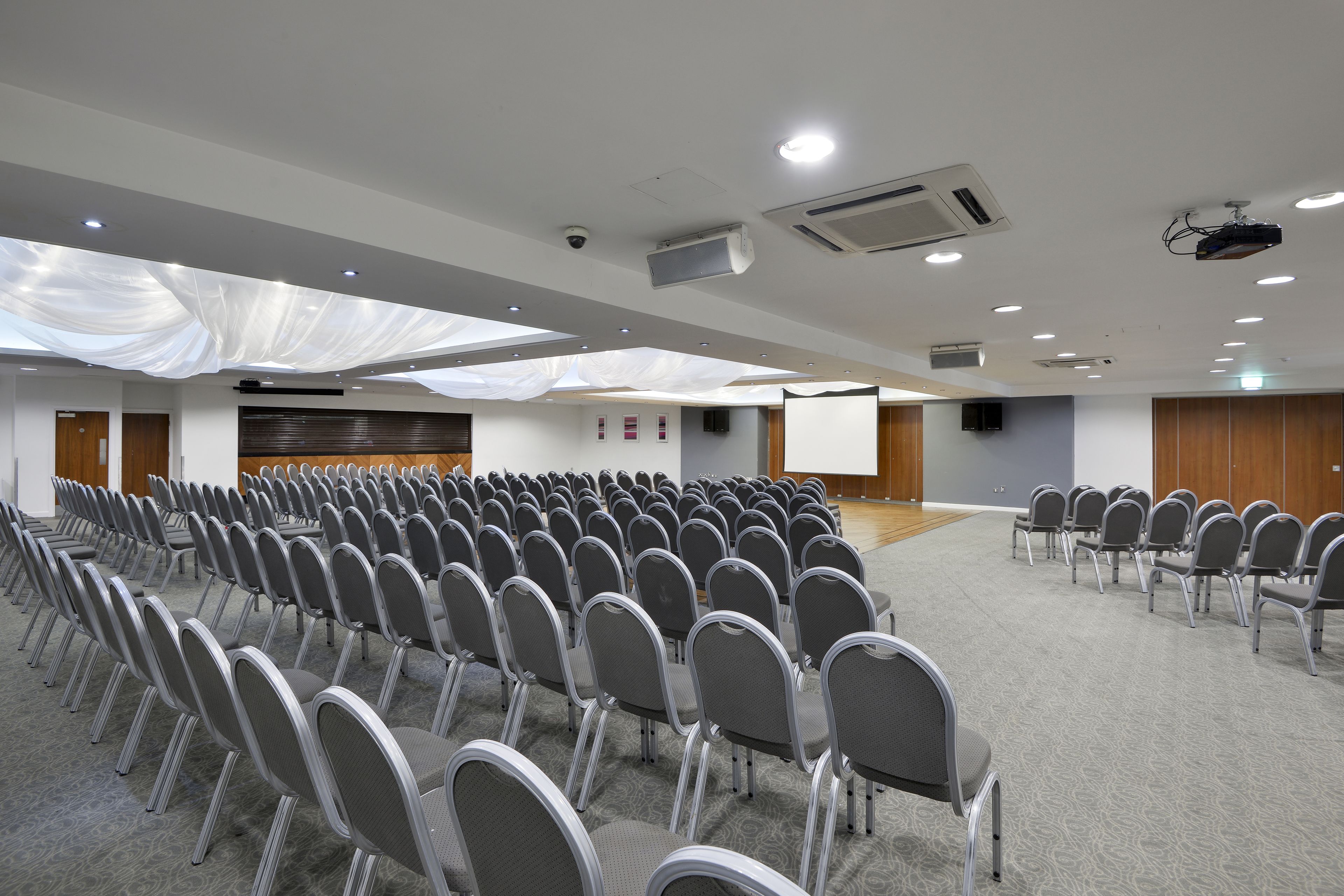 Uma sala de conferências com fileiras de cadeiras, um palco com um projetor e uma tela grande.