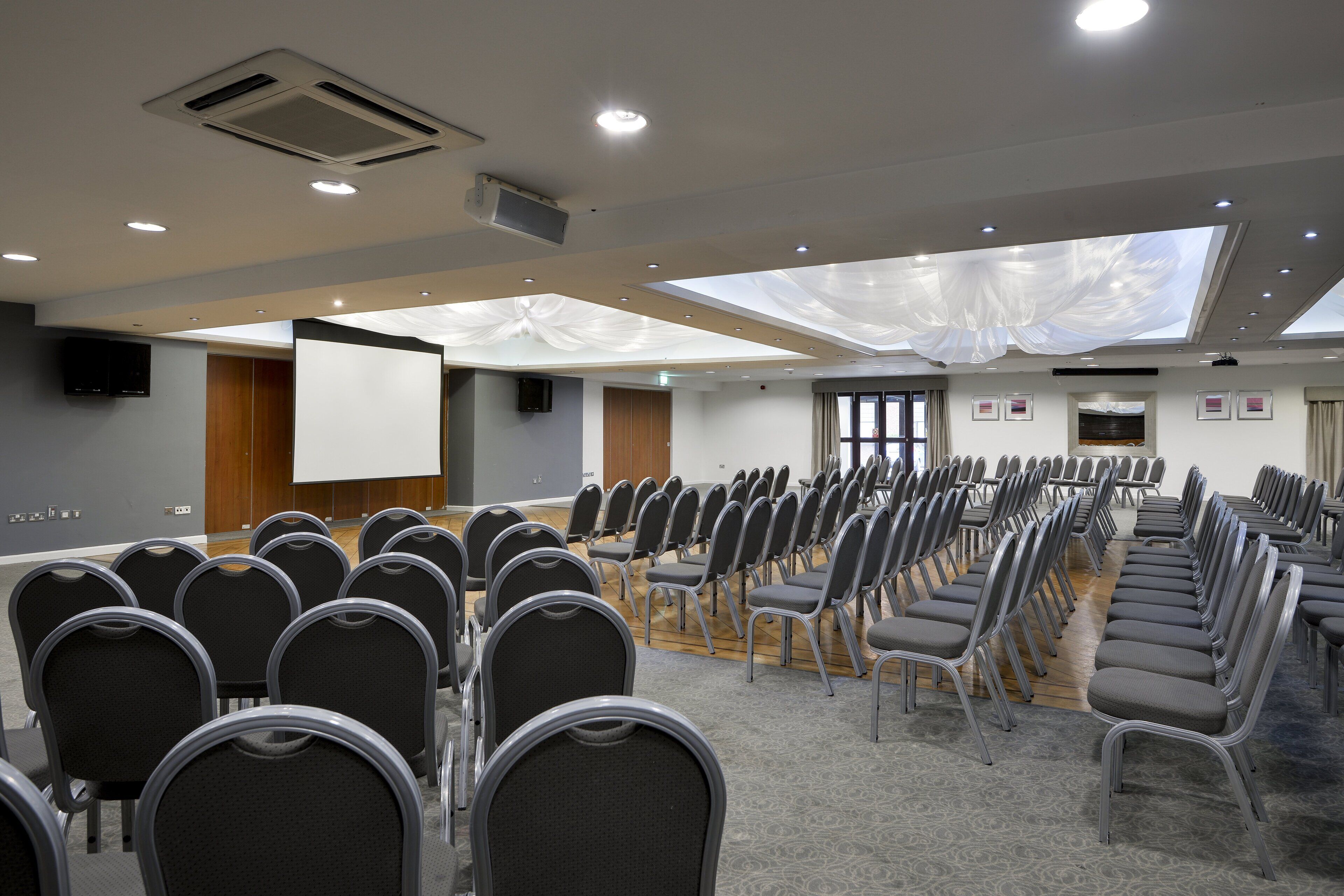 A conference room with rows of chairs, a projector screen, and a large window with a view of the sky.