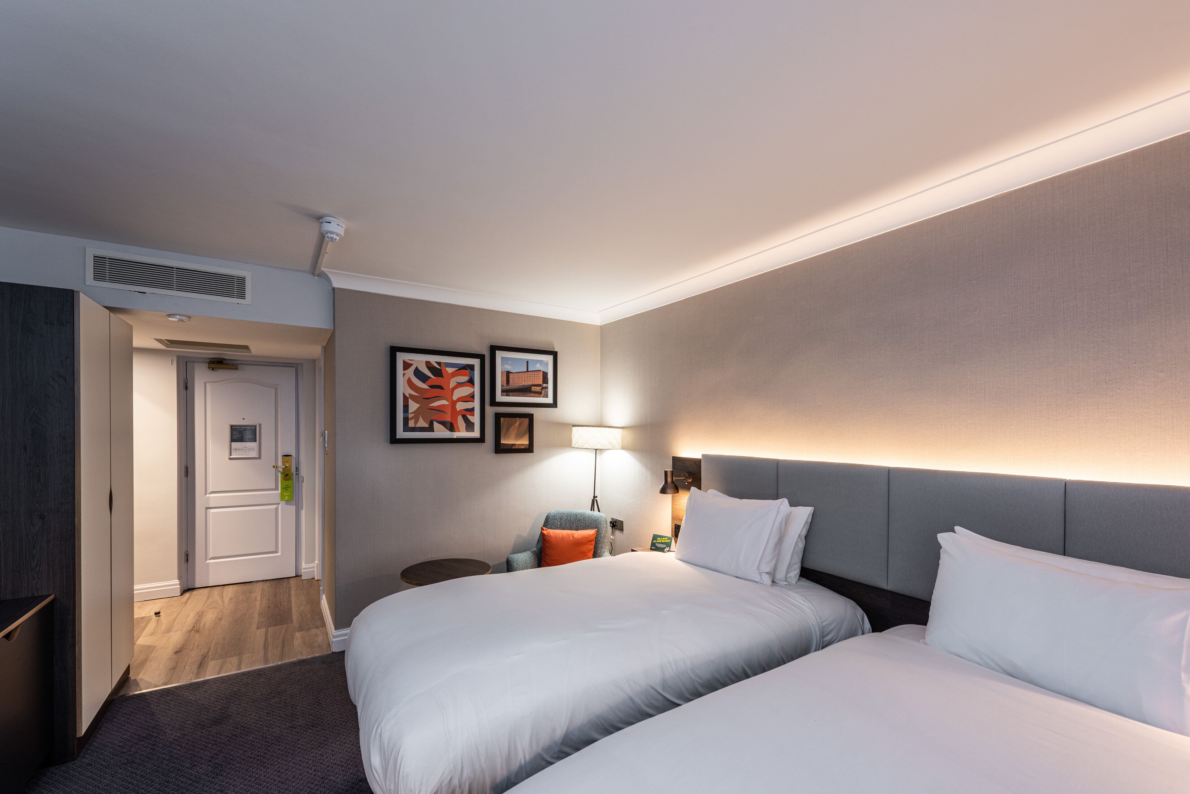 A hotel room with two beds, a desk, a chair, a lamp, and artwork on the wall.