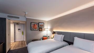 A hotel room with two beds, a desk, a chair, a lamp, and artwork on the wall.