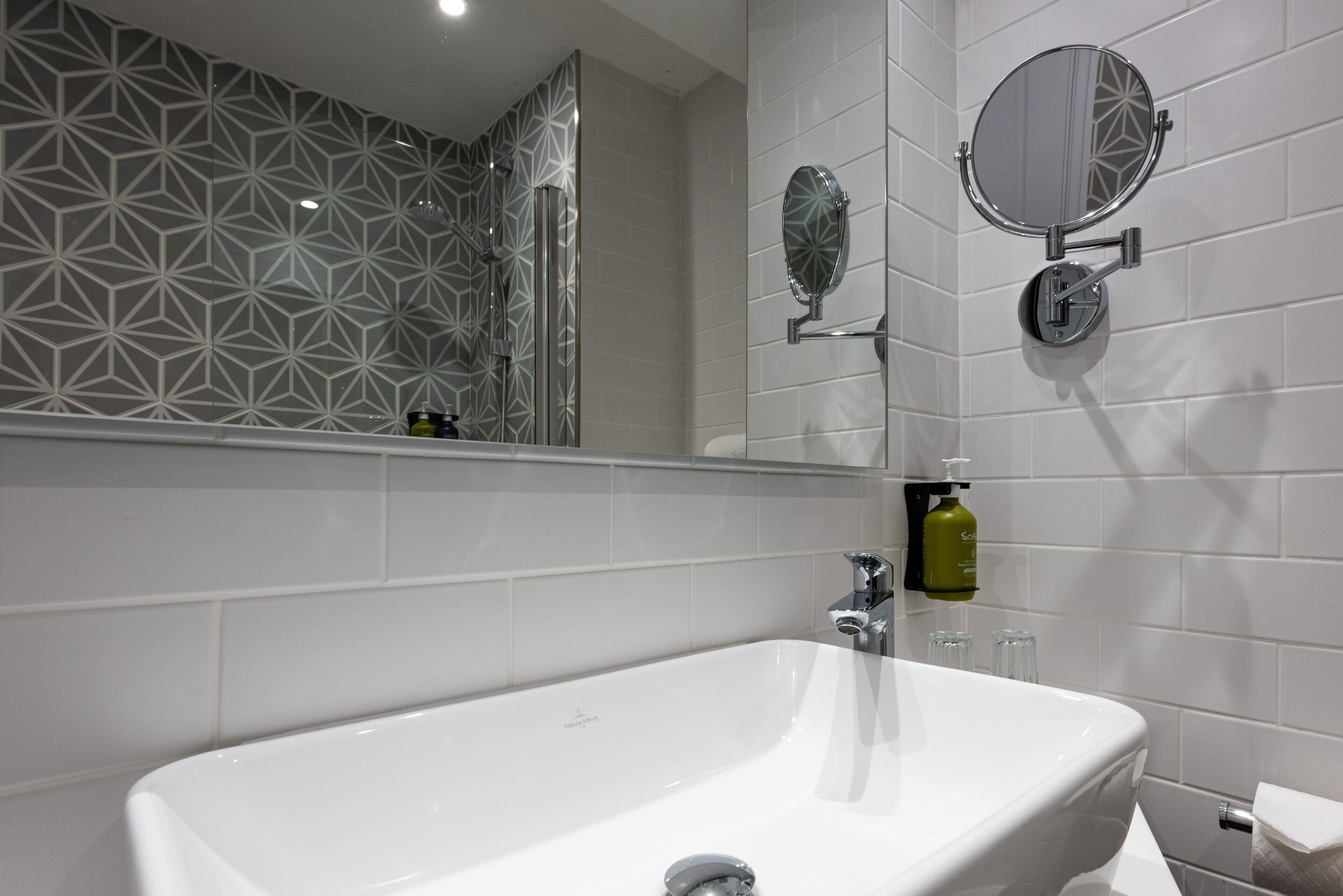 A modern bathroom with a large white sink, two wall-mounted mirrors, and a geometric patterned shower area.