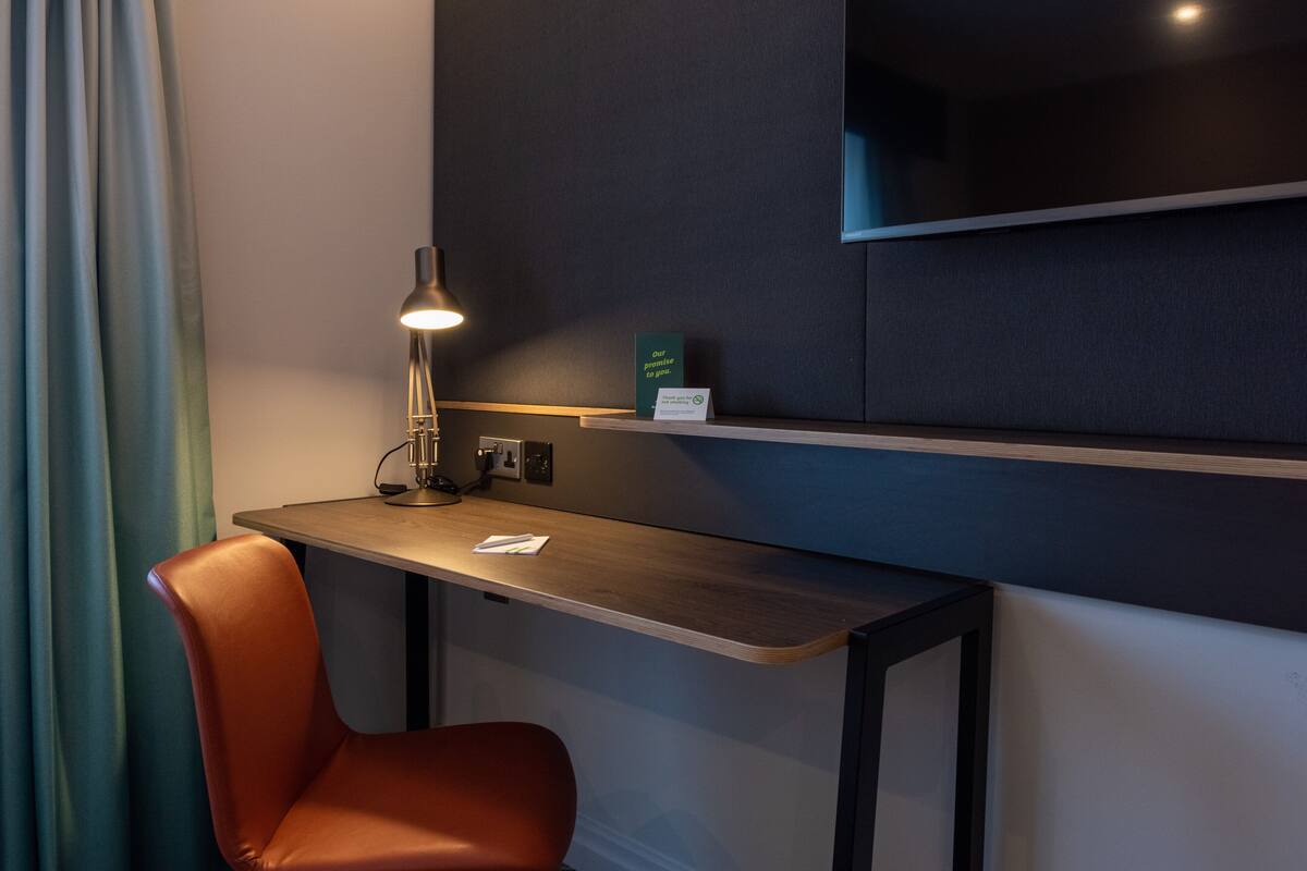 A modern hotel room with a wooden desk, a single orange chair, a desk lamp, and a flat-screen TV mounted on the wall.