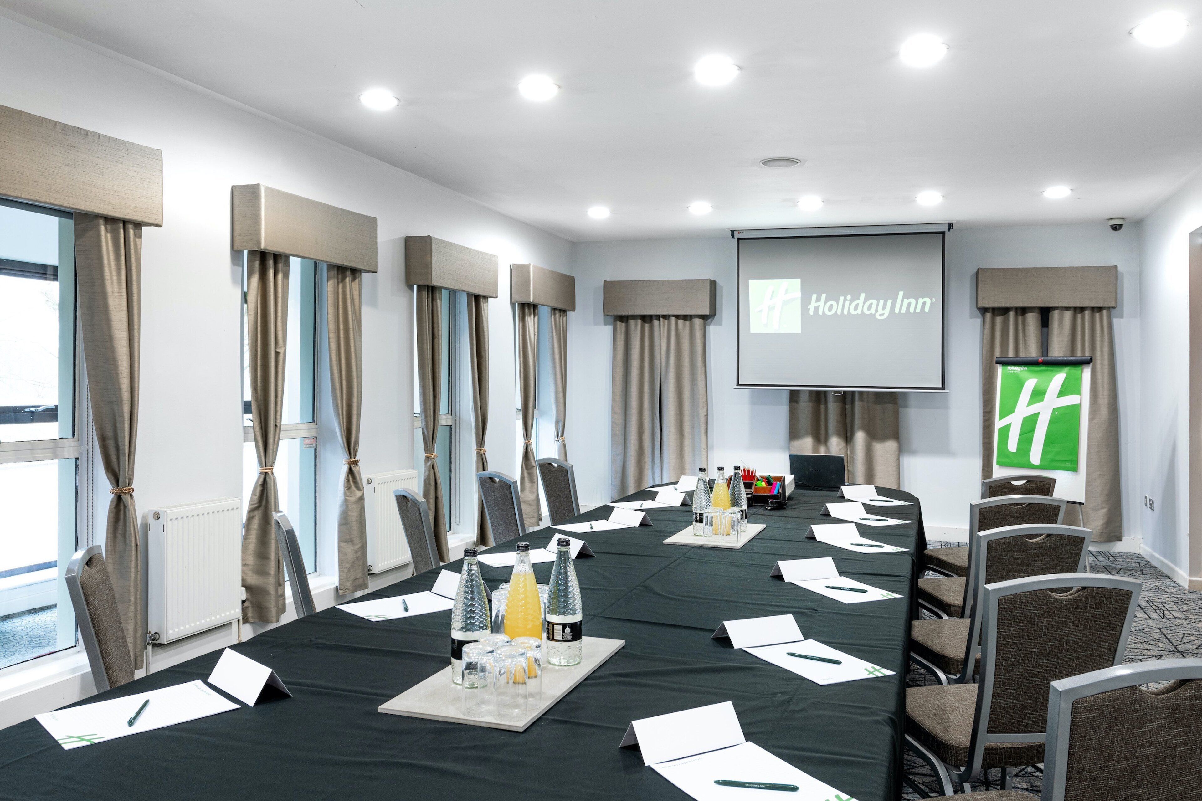 A conference room with a long table, chairs, a projector screen, and a large window with curtains.