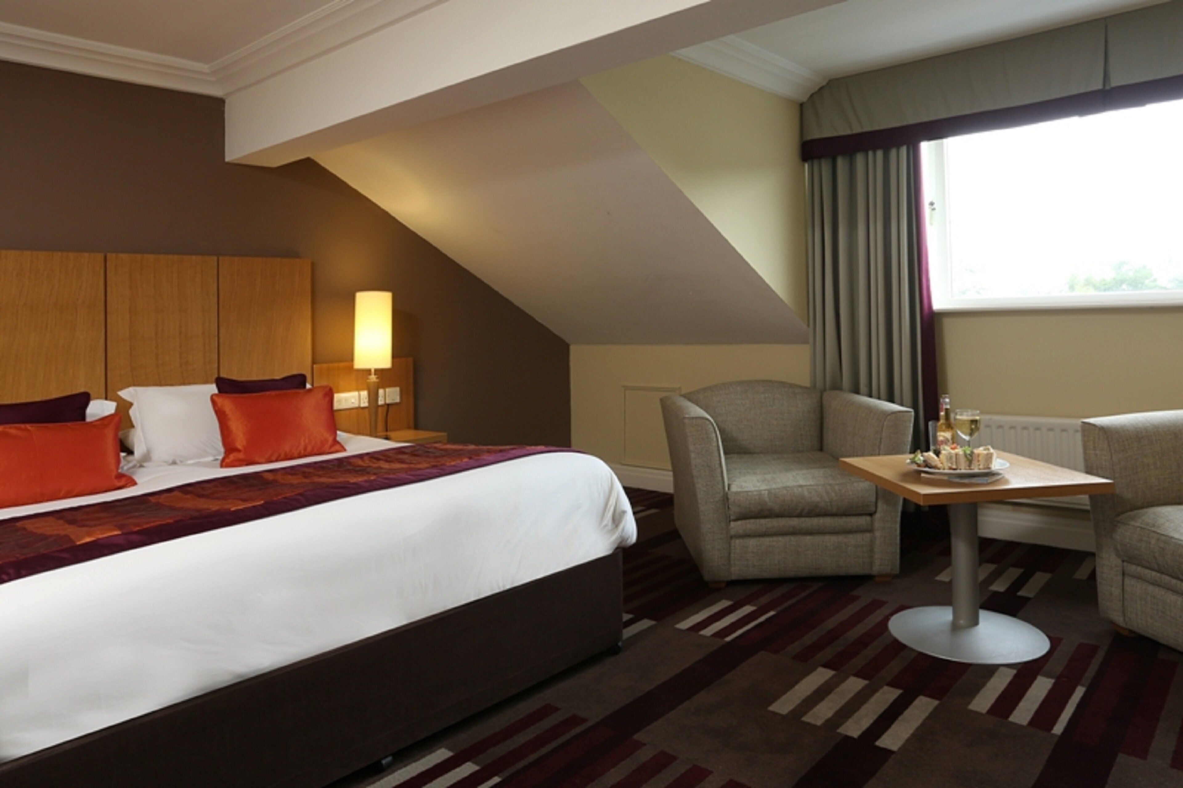 A hotel room with a large bed, two armchairs, a small table with a bottle and glasses, and a window with curtains.