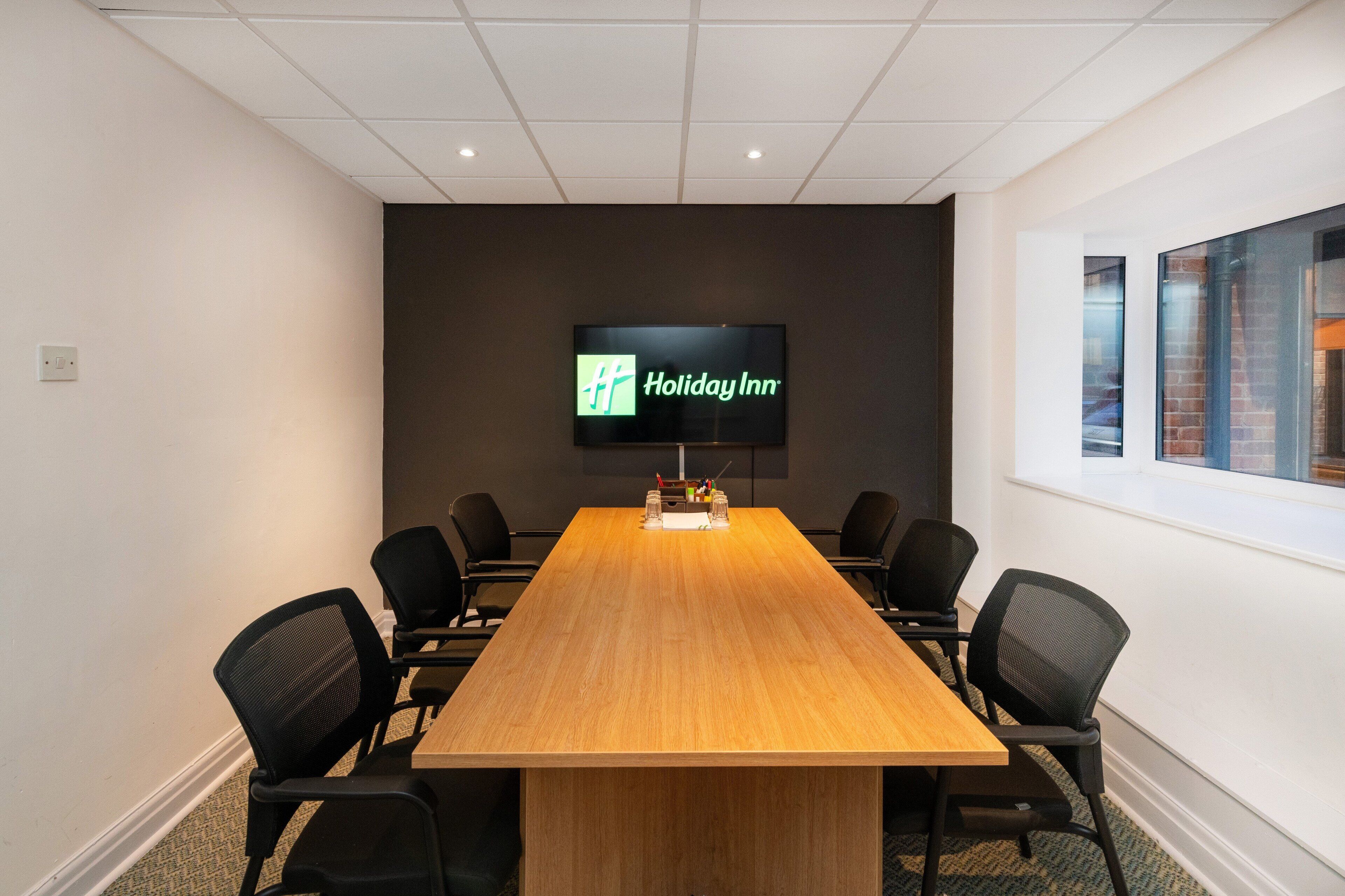 Une salle de conférence avec une longue table en bois, des chaises noires et un écran affichant le logo de Holiday Inn.