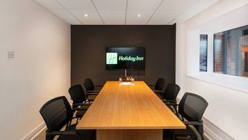 Une salle de conférence avec une longue table en bois, des chaises noires et un écran affichant le logo de Holiday Inn.