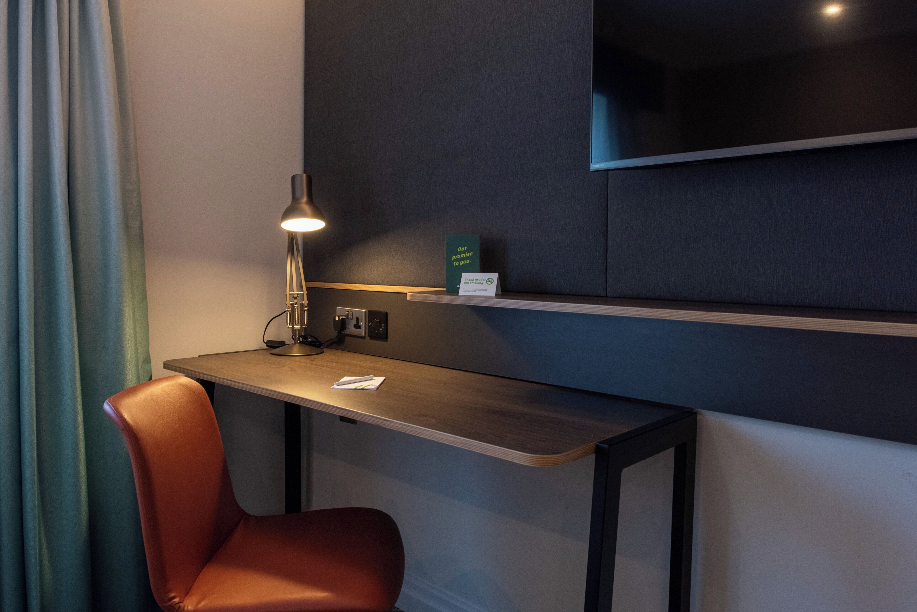 A hotel room with a desk, a chair, a lamp, a television mounted on the wall, and a sign on the wall.