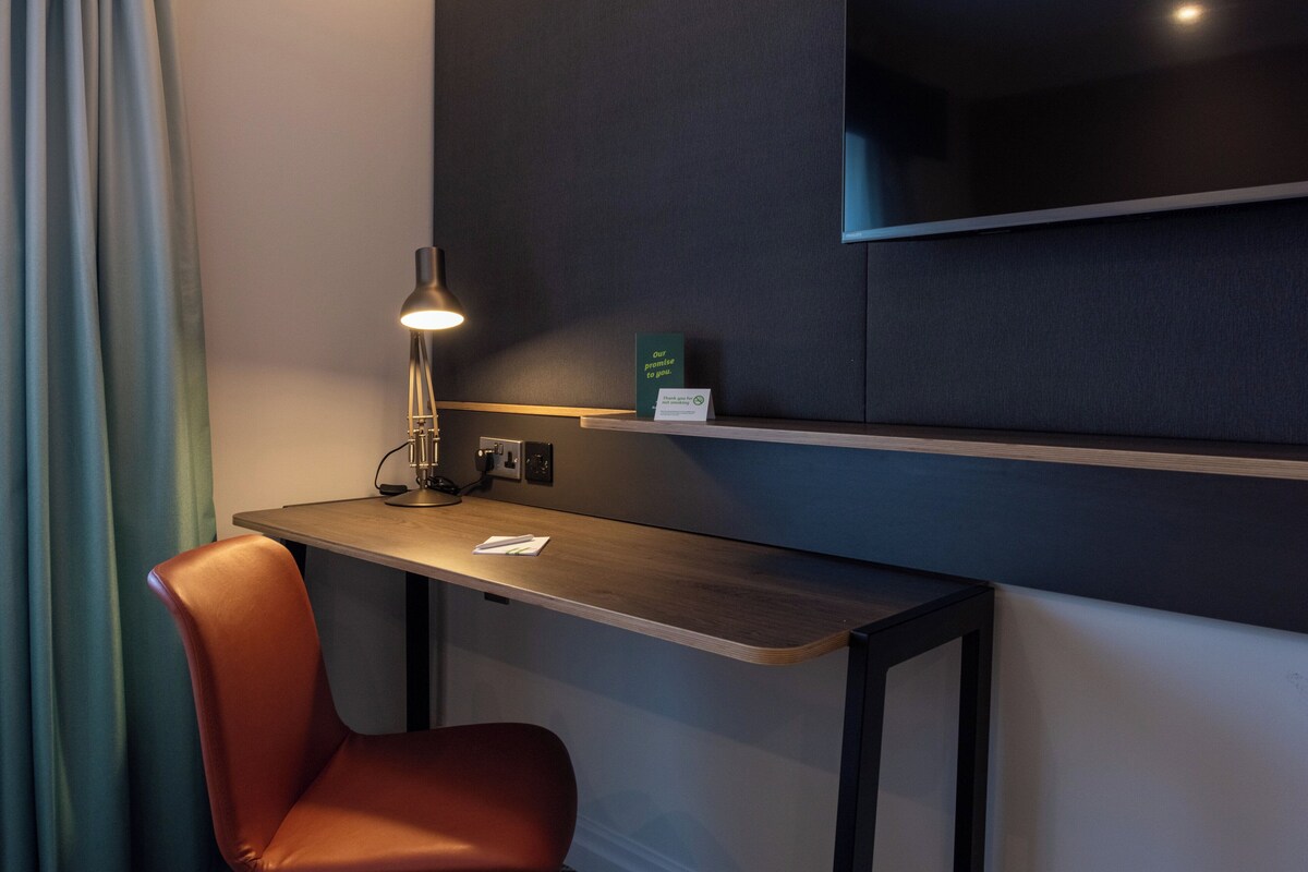 Una habitación de hotel moderna con un escritorio de madera, una sola silla naranja, una lámpara de escritorio y un televisor de pantalla plana montado en la pared.