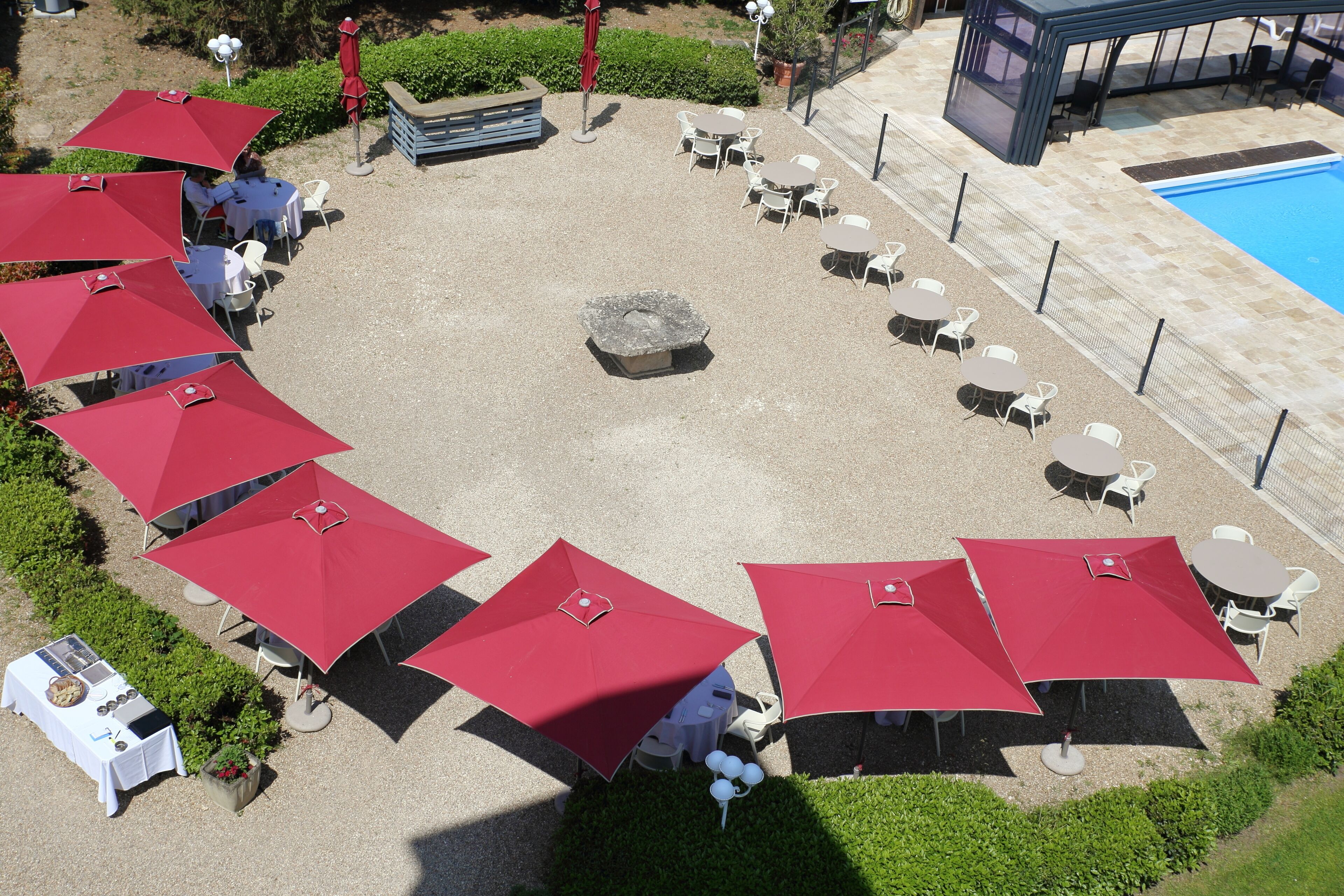 An outdoor patio area with multiple round tables and red umbrellas, surrounded by a pool and greenery.