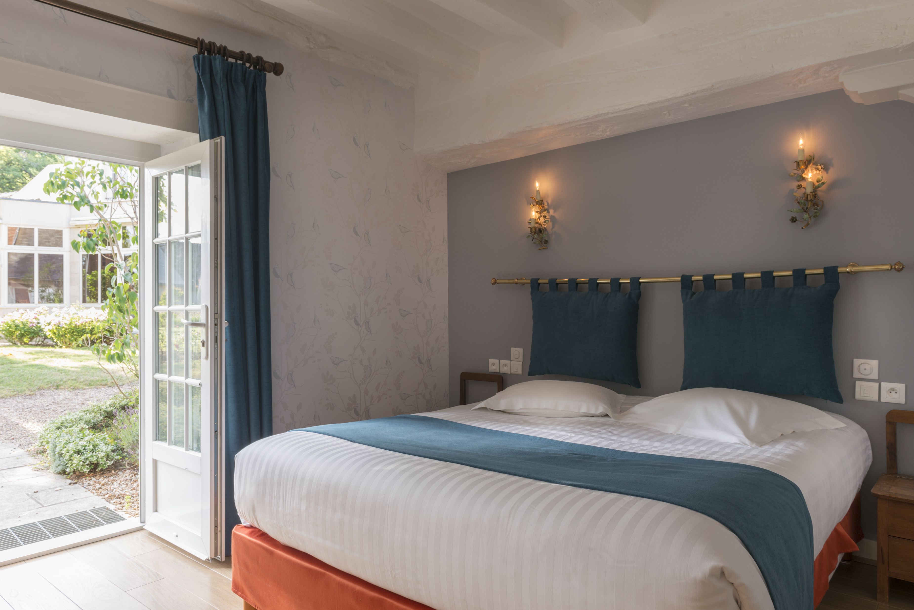 Une chambre à coucher avec un lit, une fenêtre avec des rideaux, un luminaire fixé au mur et une vue sur l’extérieur.