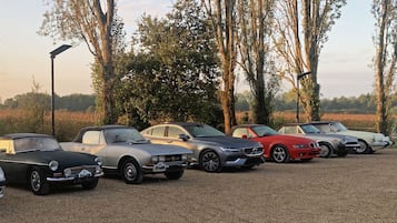 Fila de carros estacionados em um estacionamento de cascalho, com árvores e céu claro ao fundo.