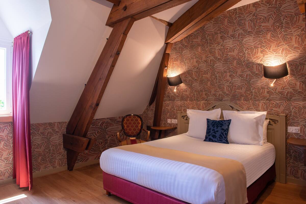 A bedroom with a sloped ceiling, a bed with white bedding, patterned wallpaper, and two wall-mounted lights.