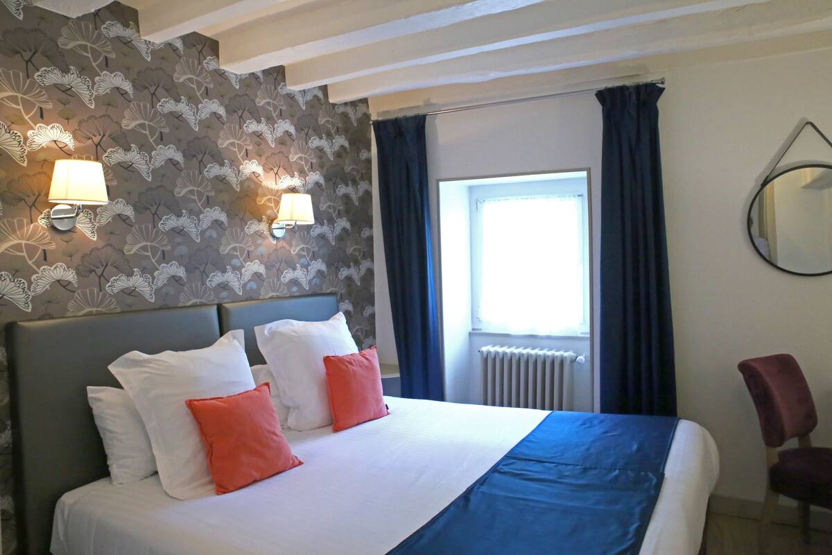 A hotel room with a bed, two wall-mounted lamps, a window with curtains, a radiator, and a chair.