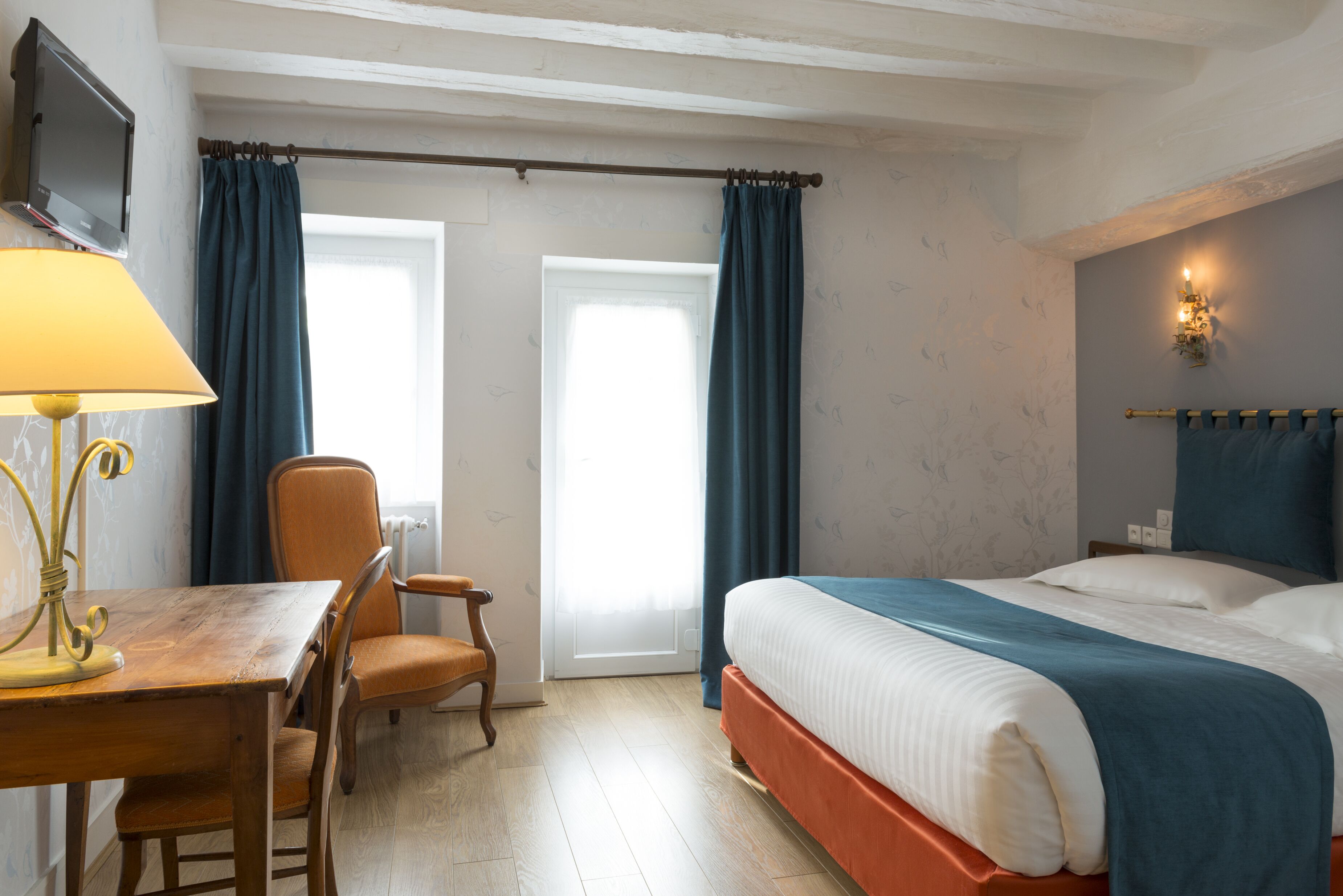 Een hotelkamer met een bed, een bureau met lamp, een stoel en een televisie aan de muur.