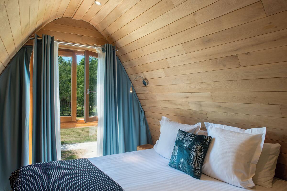 A bedroom with a wooden ceiling, a bed with white and patterned pillows, and a view of a green outdoor area through a glass door.