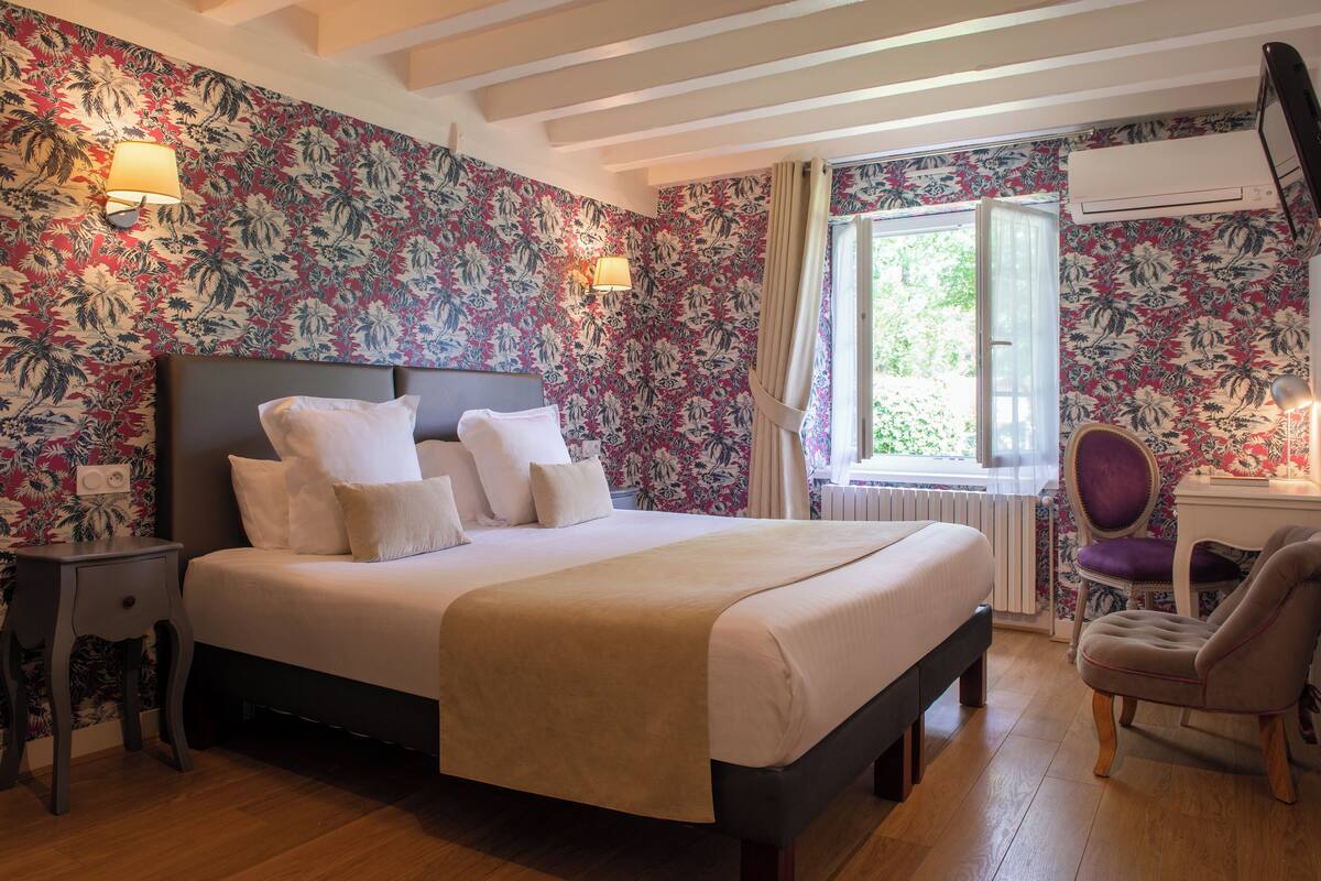 A neatly made bed with white linens and a beige throw blanket, a wooden nightstand with a lamp, a chair, and a window with curtains.
