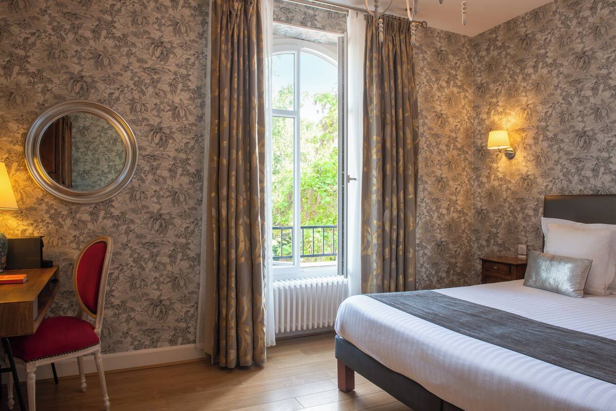 A bedroom with a bed, a desk, a chair, a mirror, and a window with curtains.