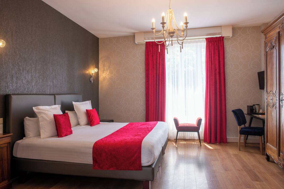 A hotel room with a large bed, a chandelier, a desk with a chair, and a wardrobe.