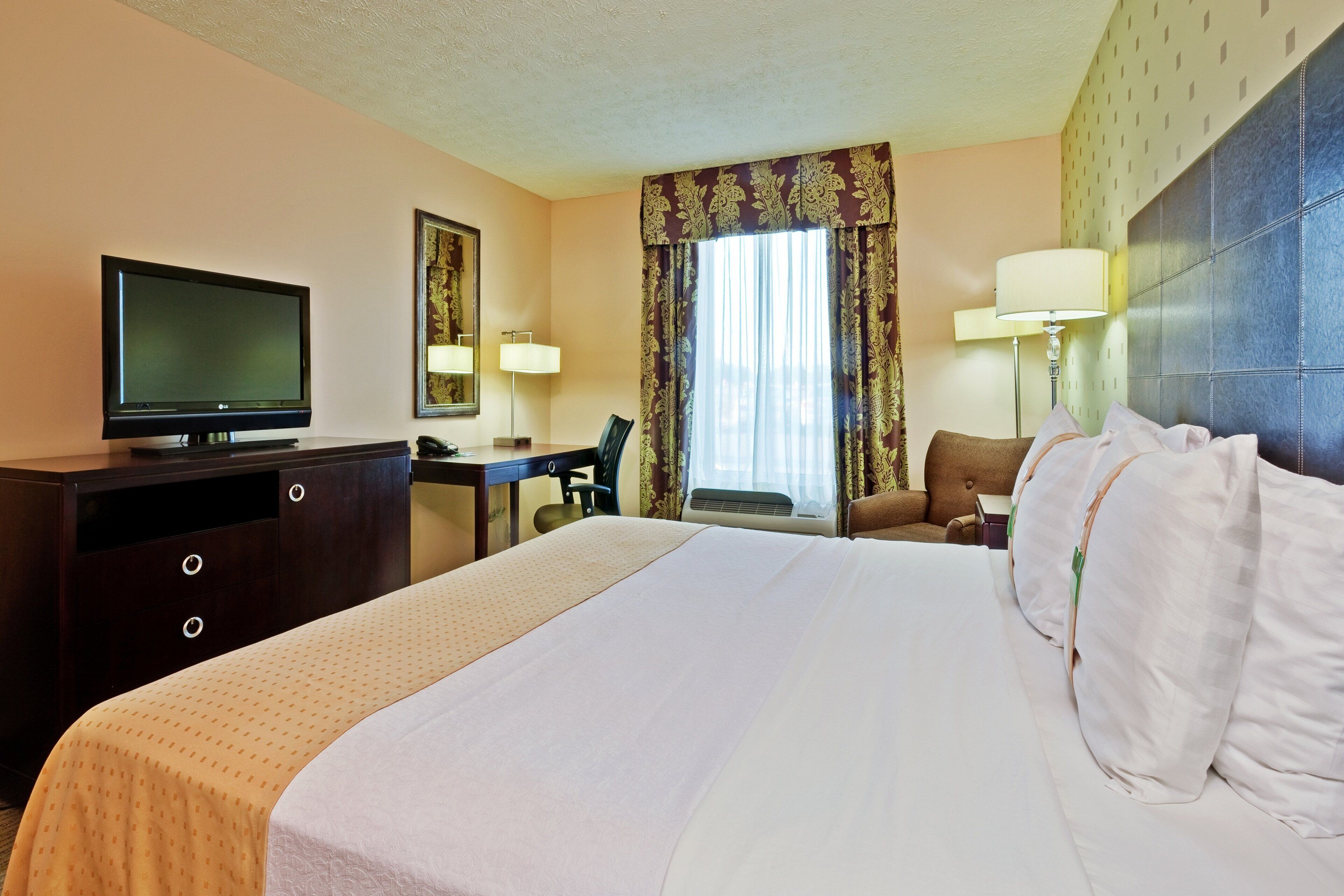 Habitación de hotel con cama, escritorio, silla, televisor y ventana con cortinas.