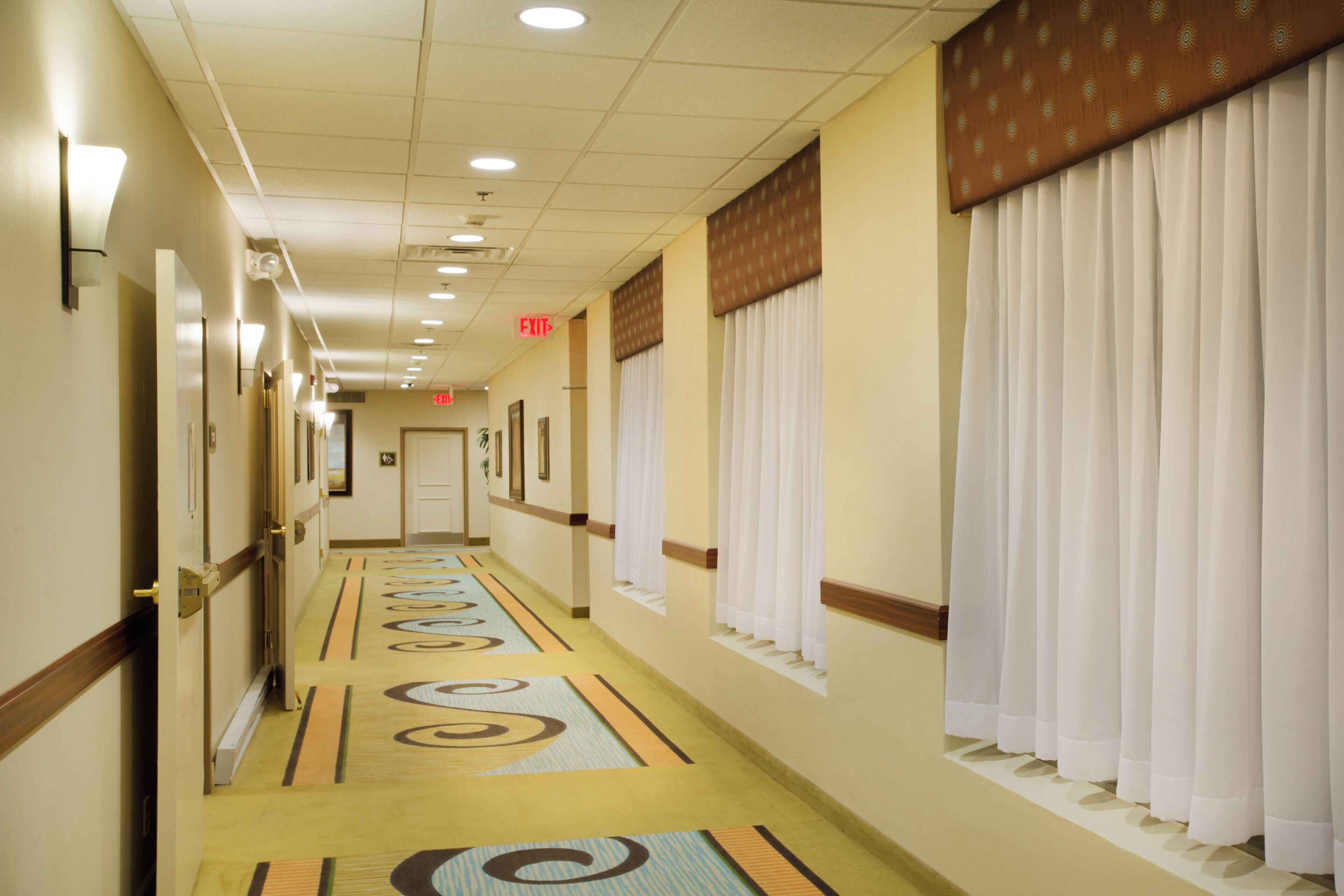 A hotel corridor with a patterned carpet, doors on the left, and windows with curtains on the right.