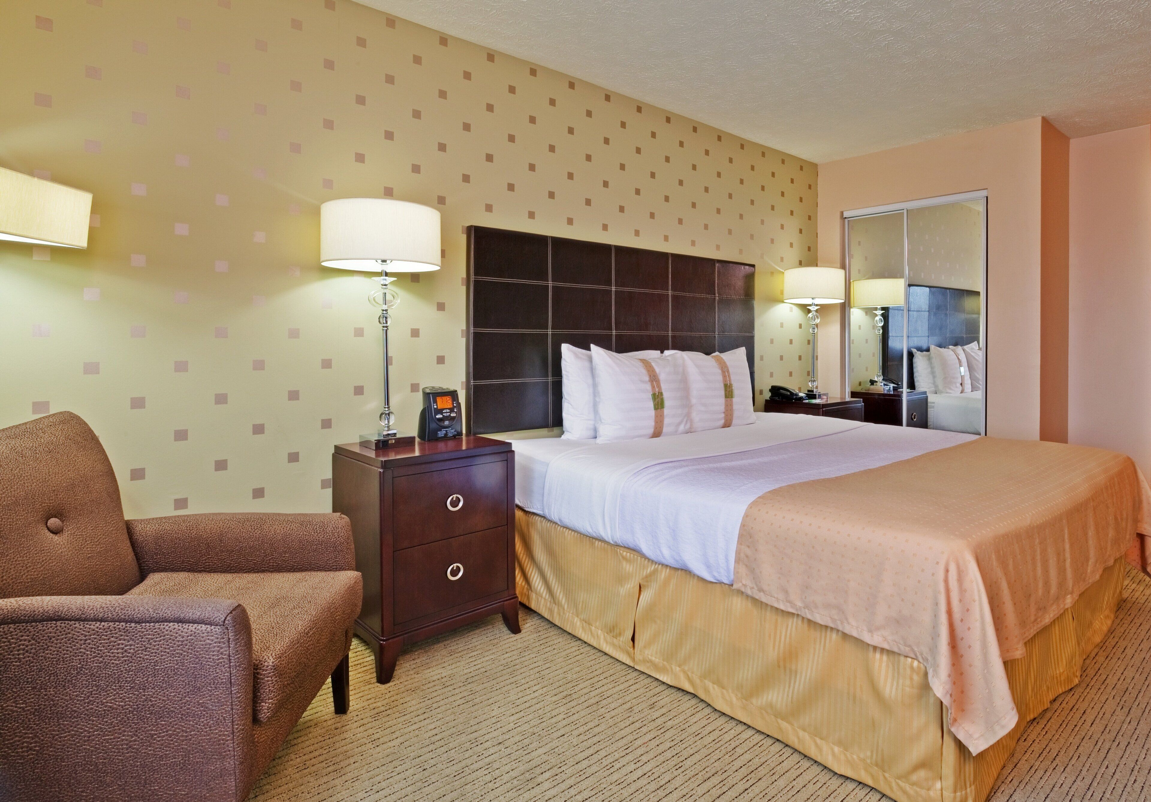 Une chambre d’hôtel avec un lit, une table de chevet, une lampe, une chaise et un miroir.