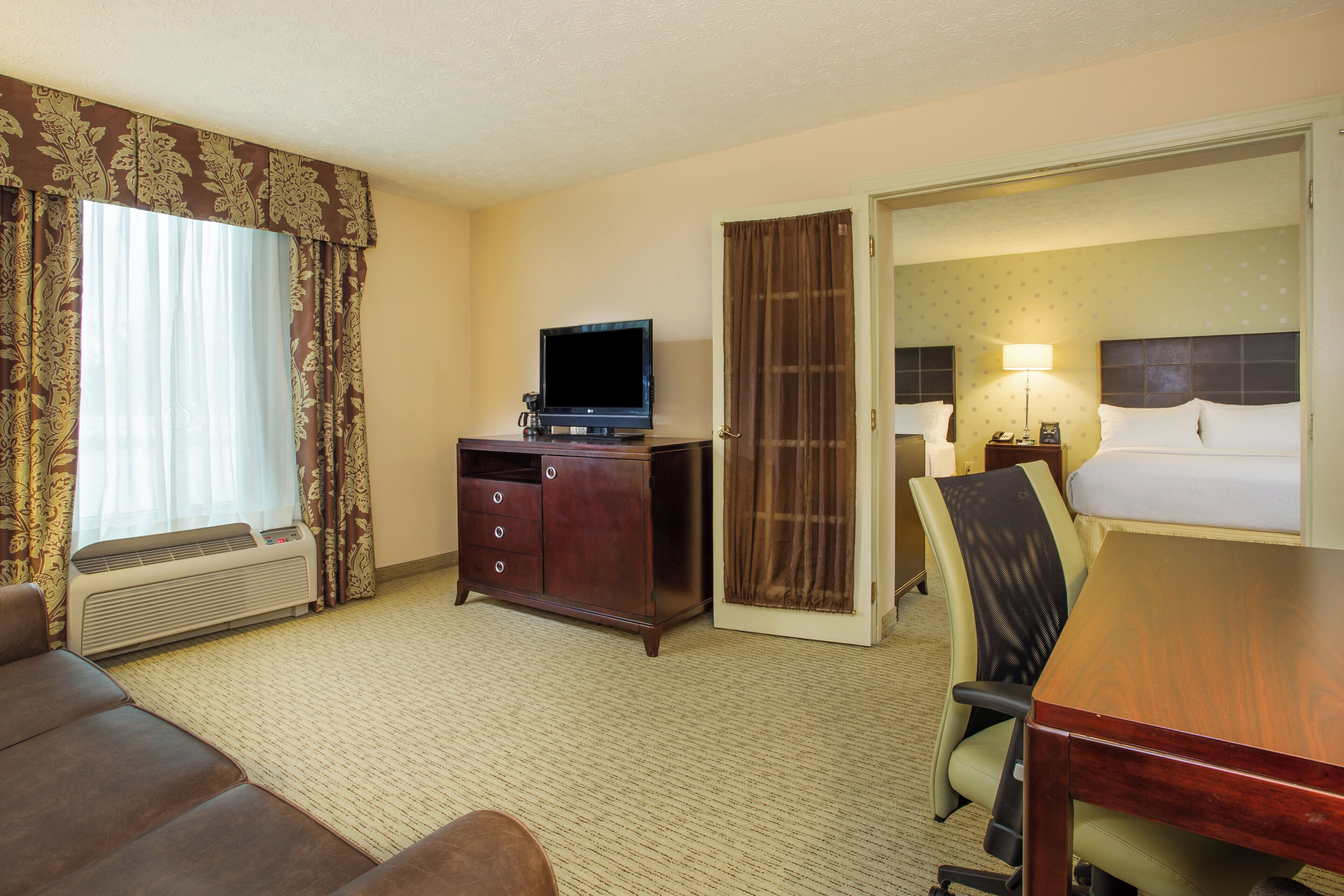 A hotel room with a flat-screen TV, a wooden dresser, a sofa, a dining table, and a separate bedroom with a bed and a nightstand.