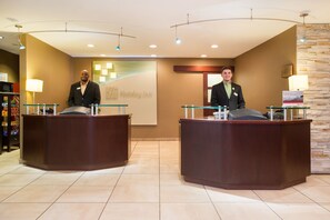Zona de recepción de hotel con dos recepcionistas en mostradores, un mostrador de recepción, una pared con un logotipo y una pared de piedra a la derecha.