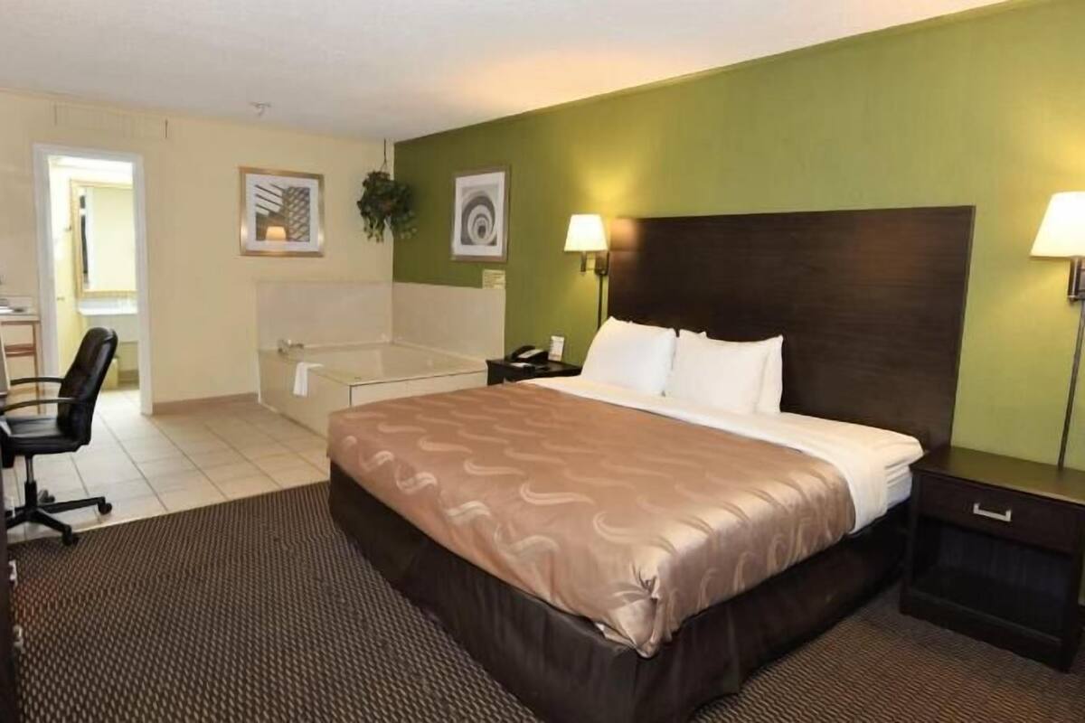 Une chambre d’hôtel avec un lit, un bureau, une chaise et une salle de bain visible à travers une porte ouverte.