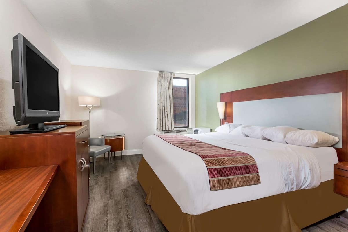Ein Hotelzimmer mit einem Bett, einem Schreibtisch mit Fernseher, einem Sessel, einer Lampe und einem Fenster mit Vorhängen.