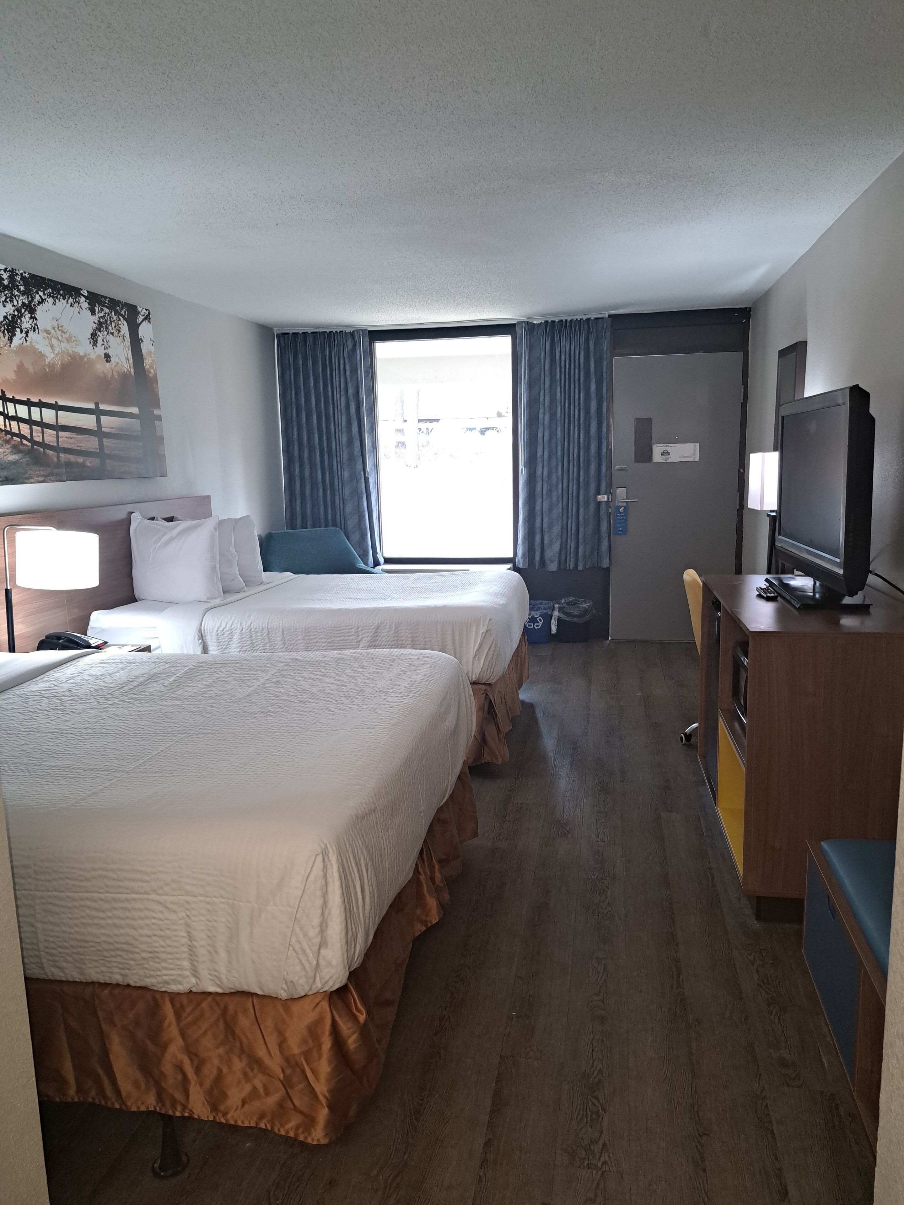 Quarto de hotel com duas camas, televisão, escrivaninha e uma janela grande com cortinas.