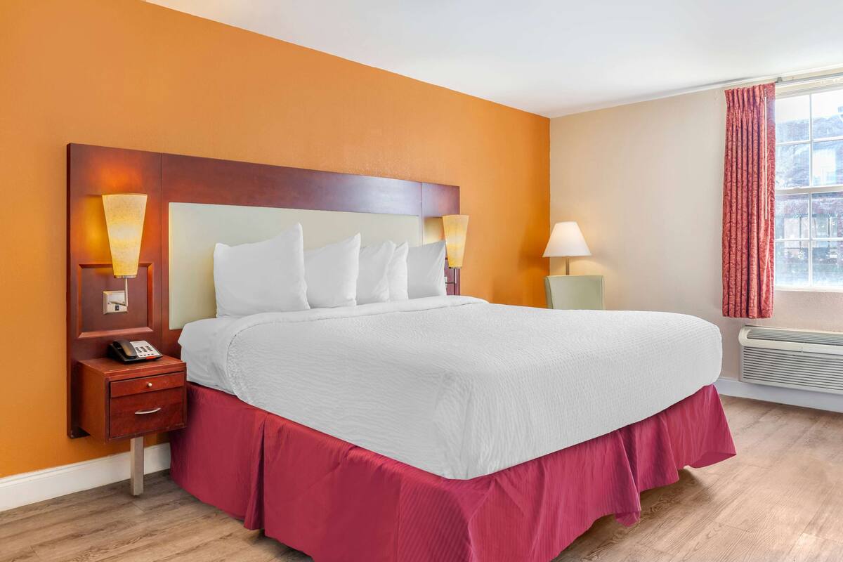 Ein Hotelzimmer mit einem großen Bett, einem Nachttisch mit Telefon, zwei Wandleuchten und einem Fenster mit Vorhängen.