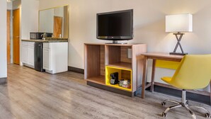 Ein Hotelzimmer mit Holzboden, einem Schreibtisch mit gelbem Stuhl, einem Fernseher, einem Spiegel und einer kleinen Küchenzeile.