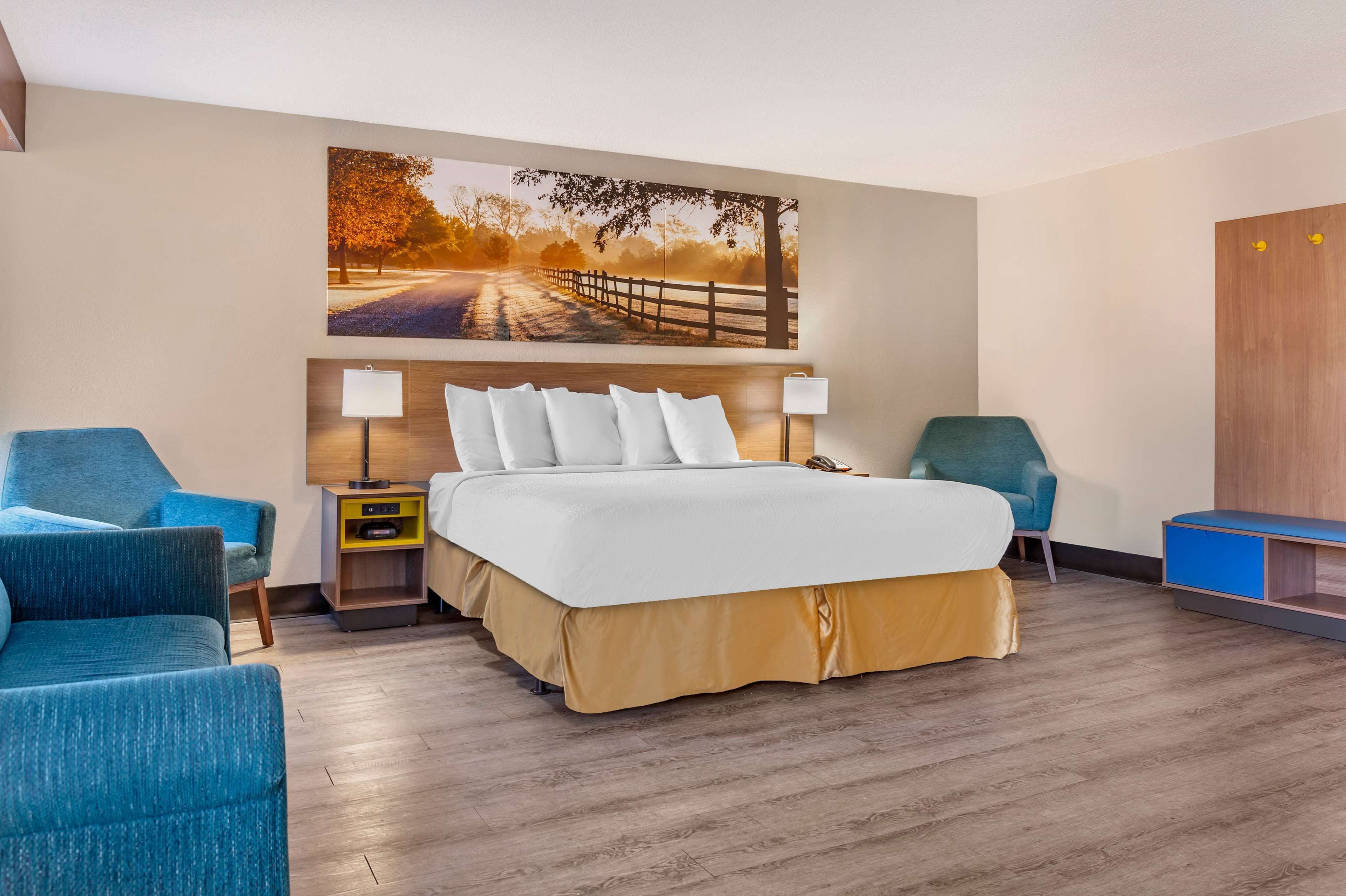 Una habitación de hotel con una cama grande, dos mesitas de noche con lámparas, un sillón azul y un sillón verde azulado, además de un cuadro grande en la pared que muestra una carretera.