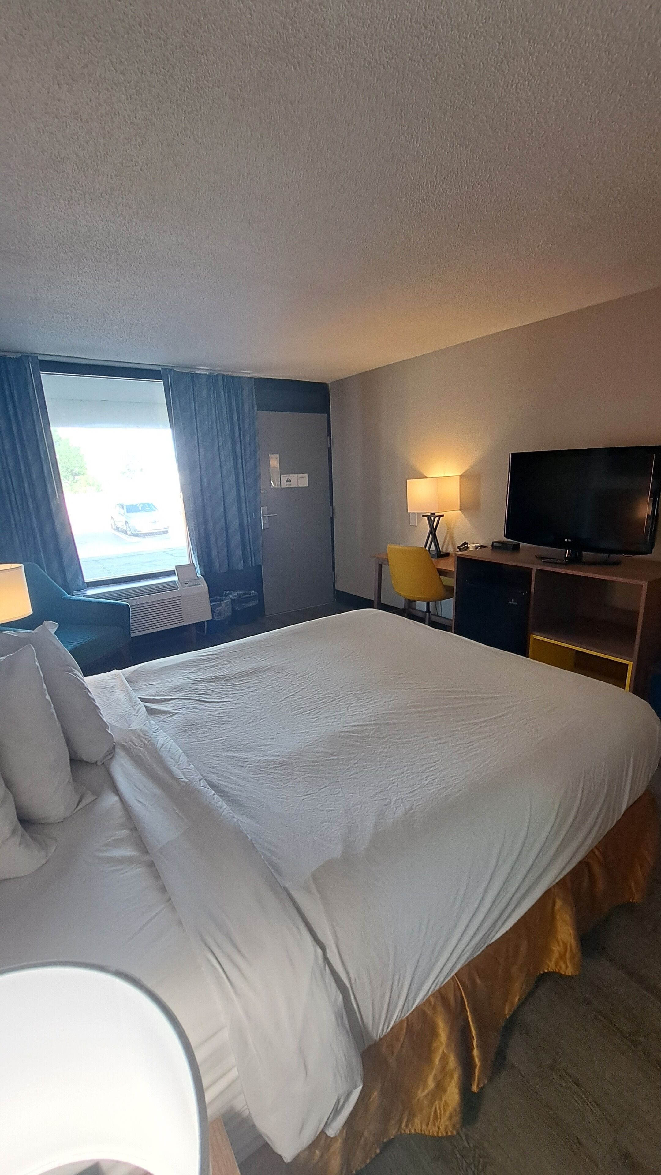 Une chambre d’hôtel avec un grand lit, un bureau avec une lampe, une télévision et une fenêtre avec des rideaux.