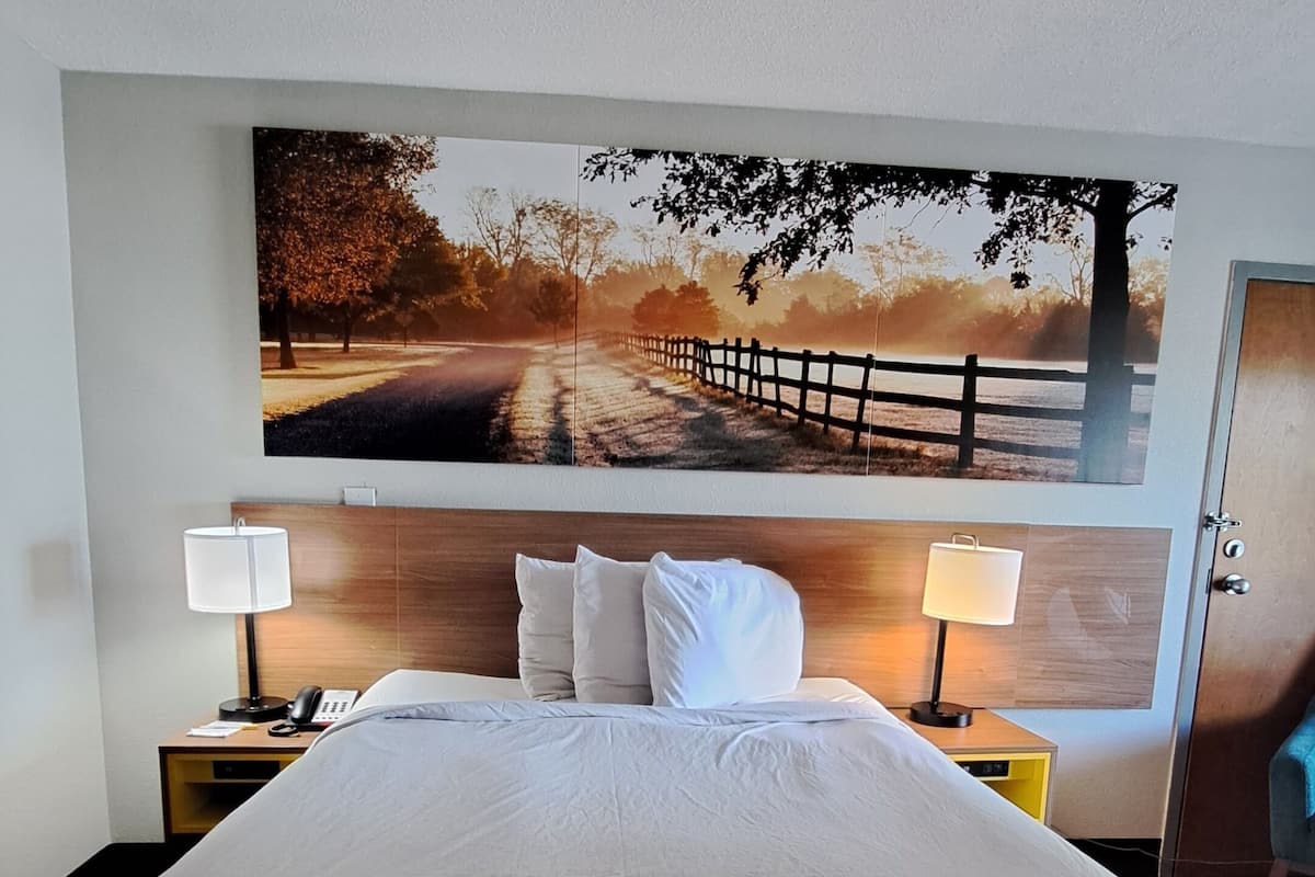 Ein Hotelzimmer mit einem großen Bett, zwei Nachttischlampen, einem Holzkopfteil und einem großen Landschaftsbild an der Wand.