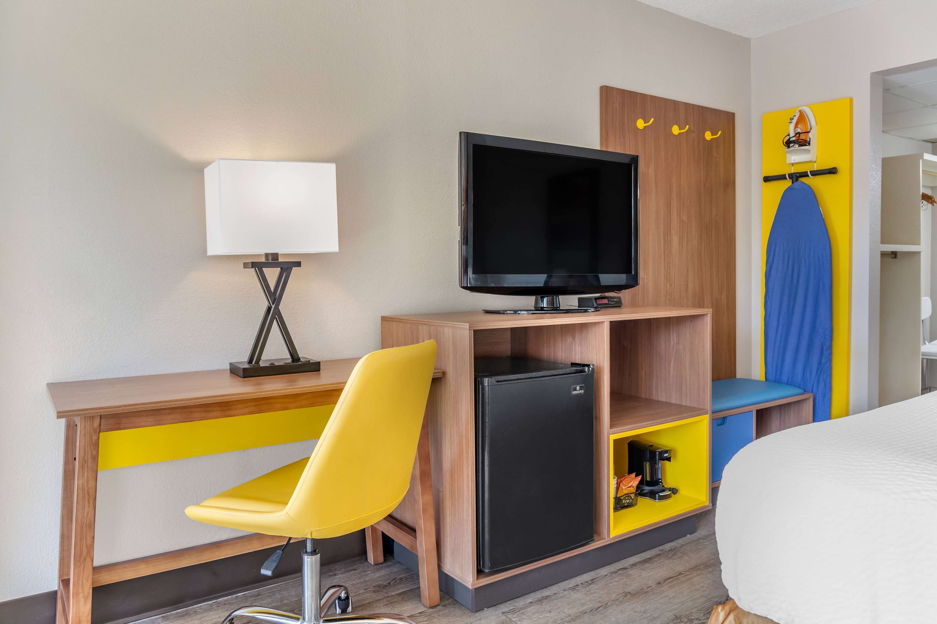 A hotel room with a wooden desk, a yellow chair, a black refrigerator, a television, and a bed.