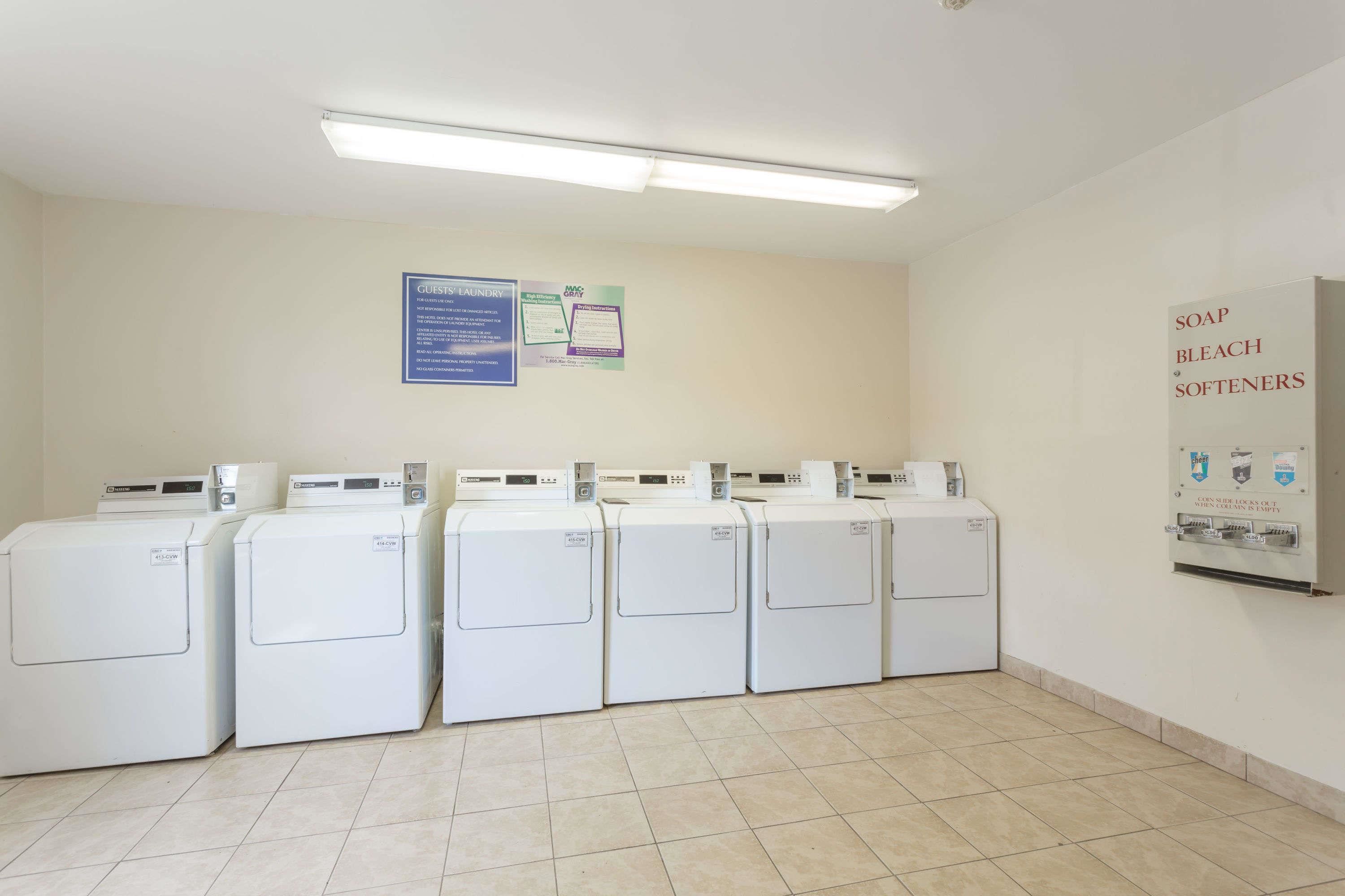 A laundry room with five front-loading washers and dryers, a wall-mounted soap and bleach dispenser, and a bulletin board with laundry instructions.