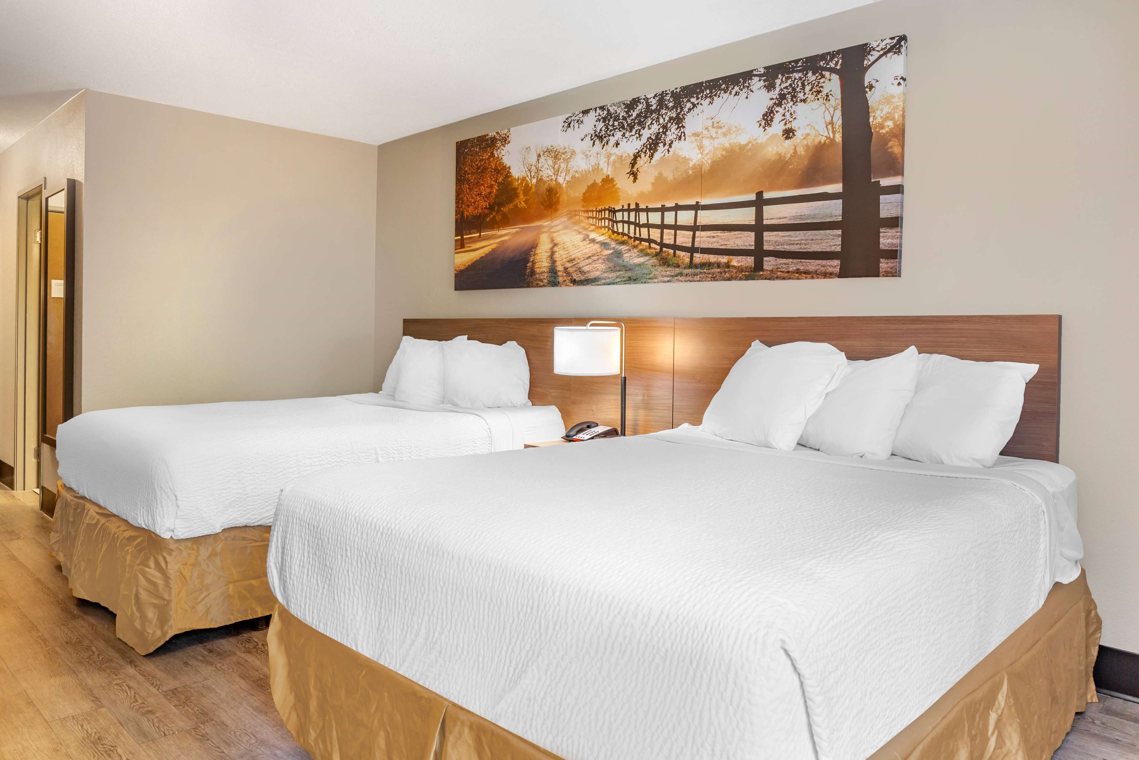 A hotel room with two beds, a large painting of a sunset road, and a nightstand with a lamp.