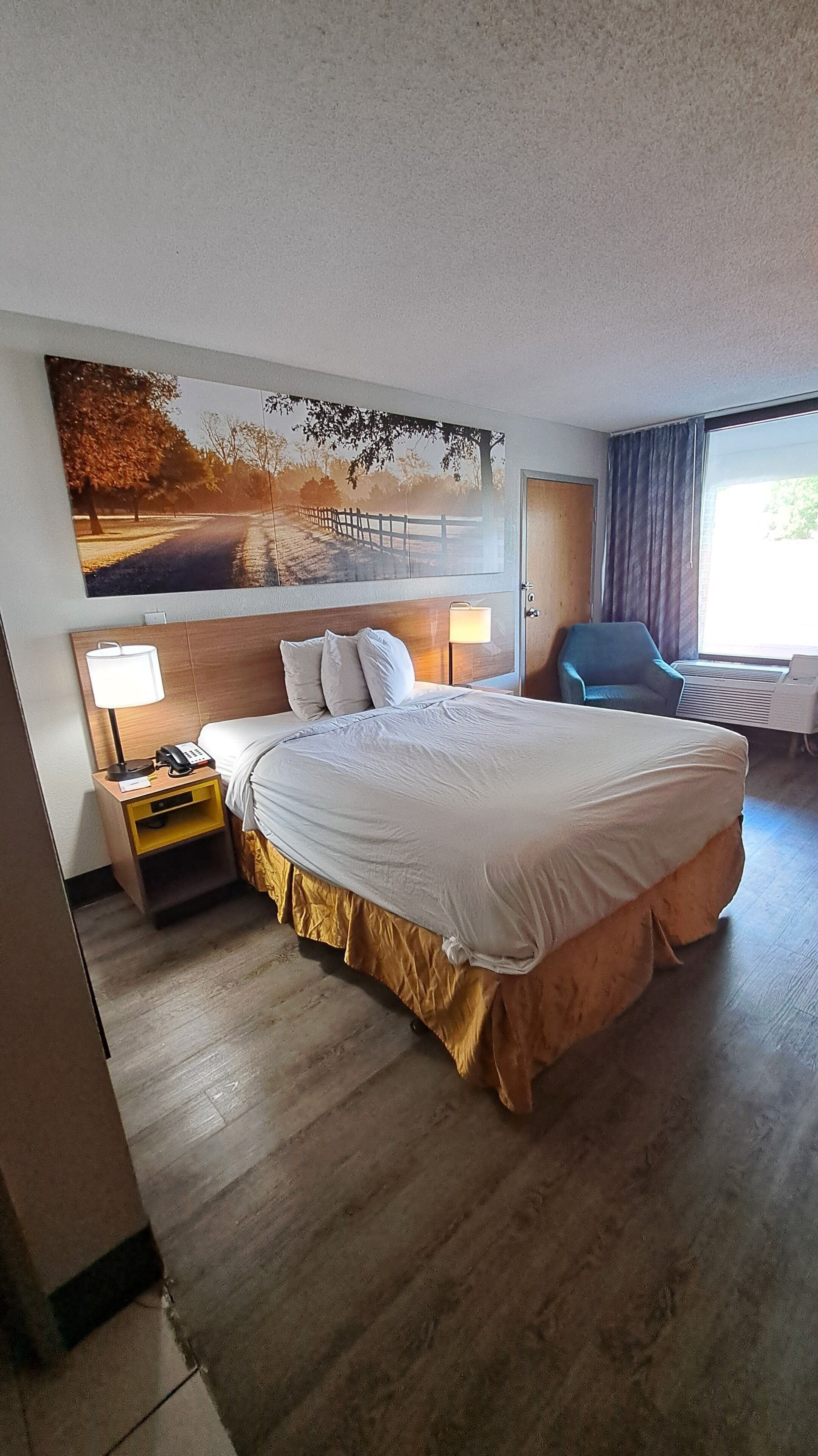 A hotel room with a large bed, a nightstand with a lamp, a chair, and a wall-mounted TV.