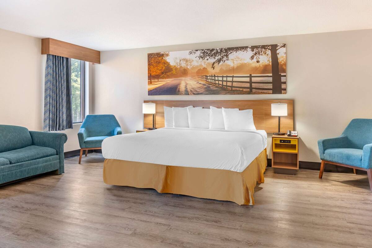Ein Hotelzimmer mit einem großen Bett, zwei Sesseln, einem Nachttisch und einem Gemälde einer malerischen Straße.